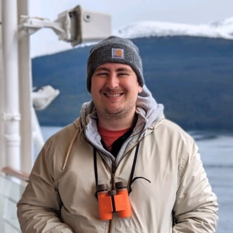 Smiling man wearing a gray beanie, beige jacket, and orange binoculars around his neck standing outdoors near water and mountains.