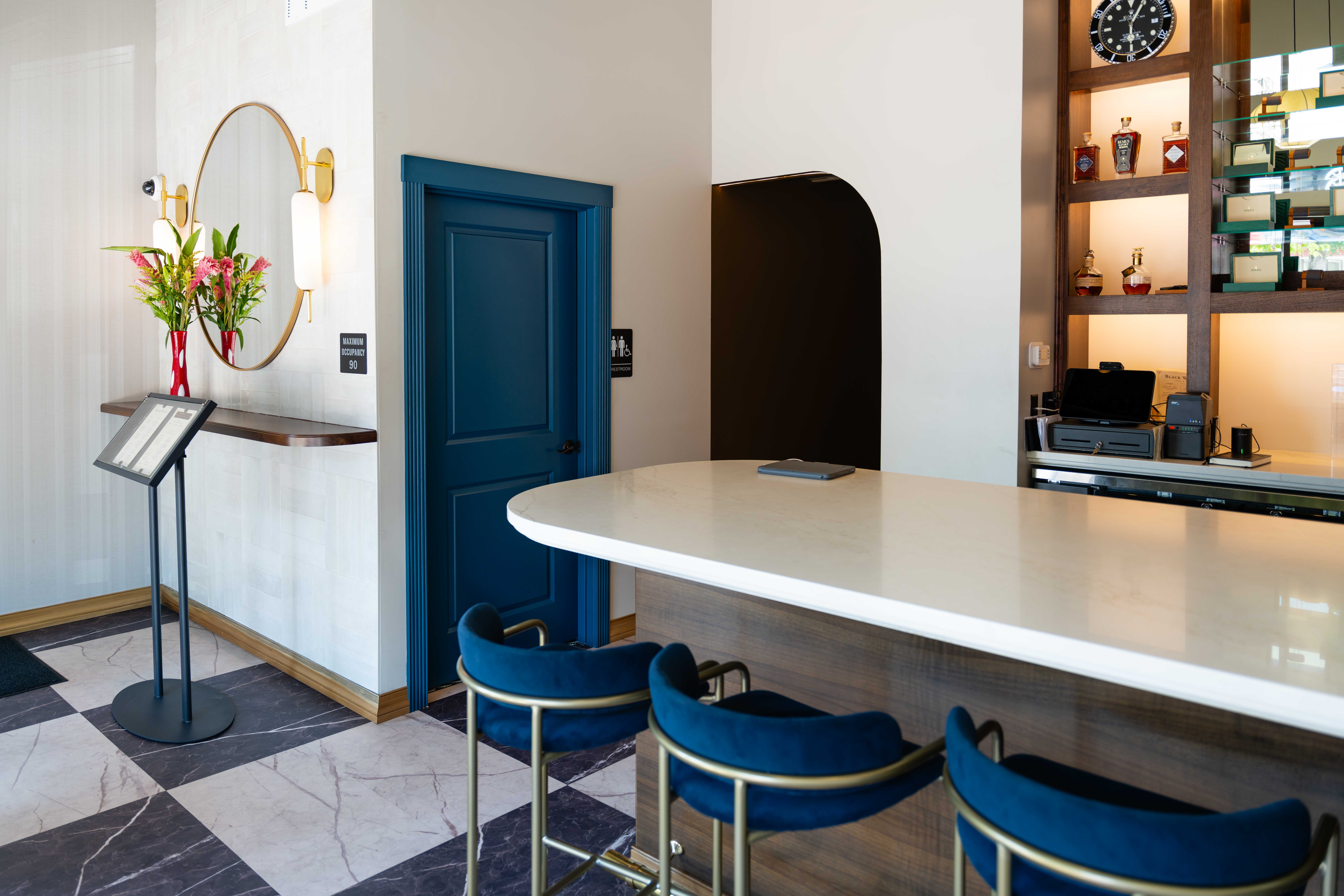 Entry with a navy door, round gold mirror, marble checkered floor and a curved white counter with blue velvet stools