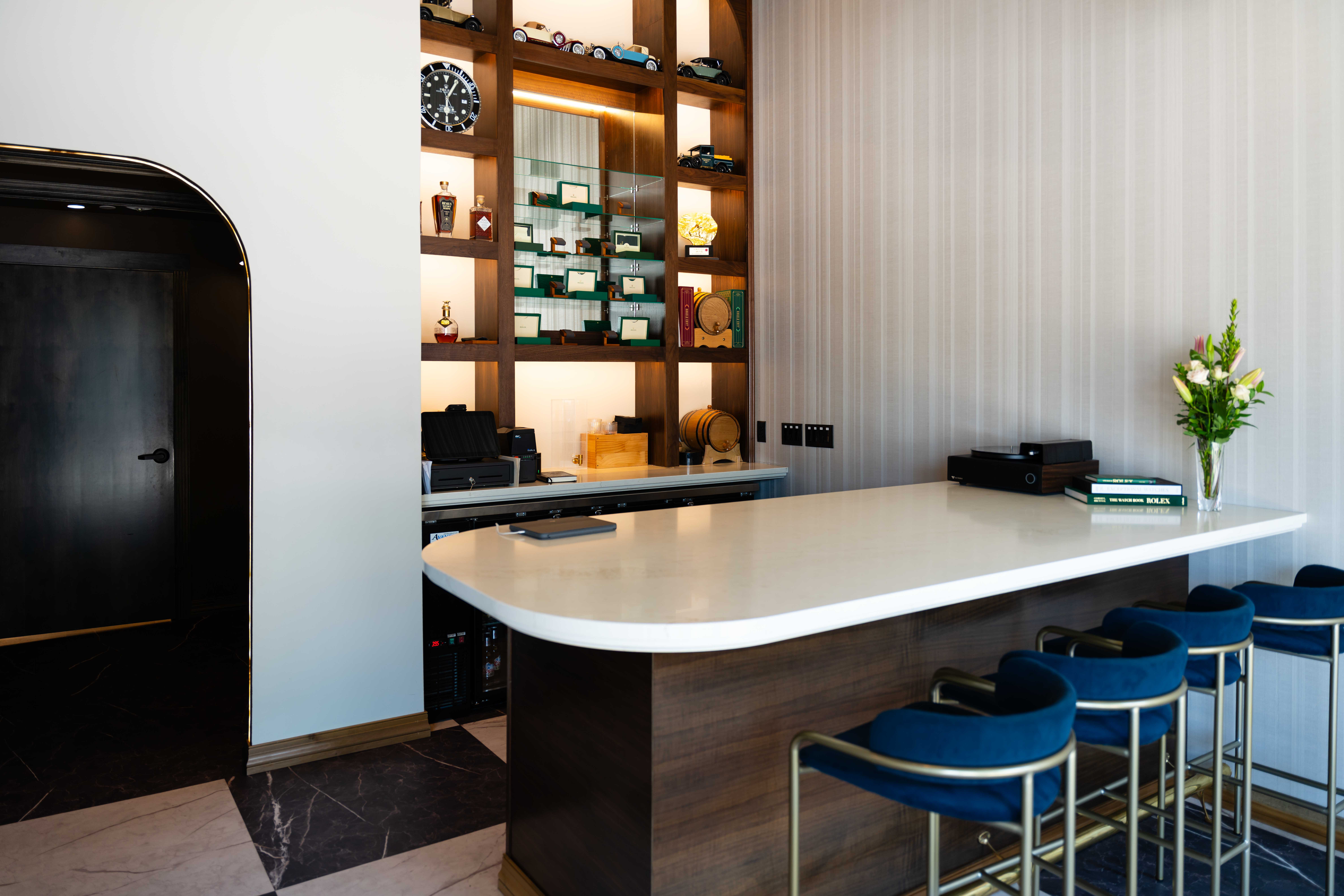 White quartz bar counter on a dark wood base with blue velvet stools in front of lit walnut display shelving
