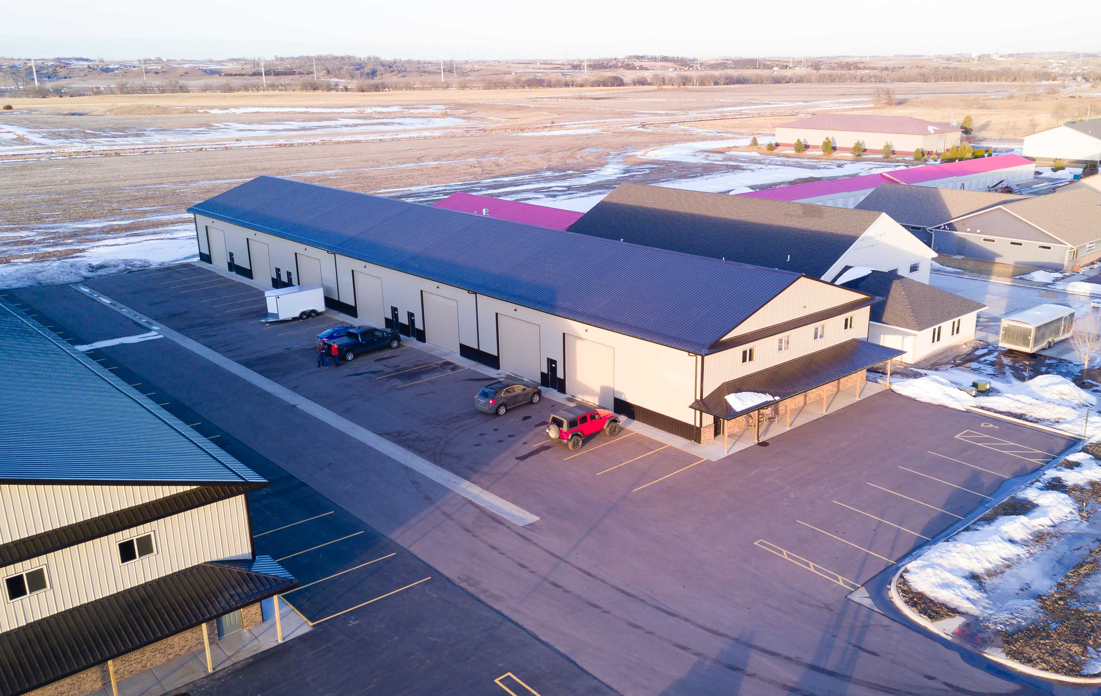 Aerial view of a tan steel shop condo building with dark trim and overhead doors in a winter parking lot