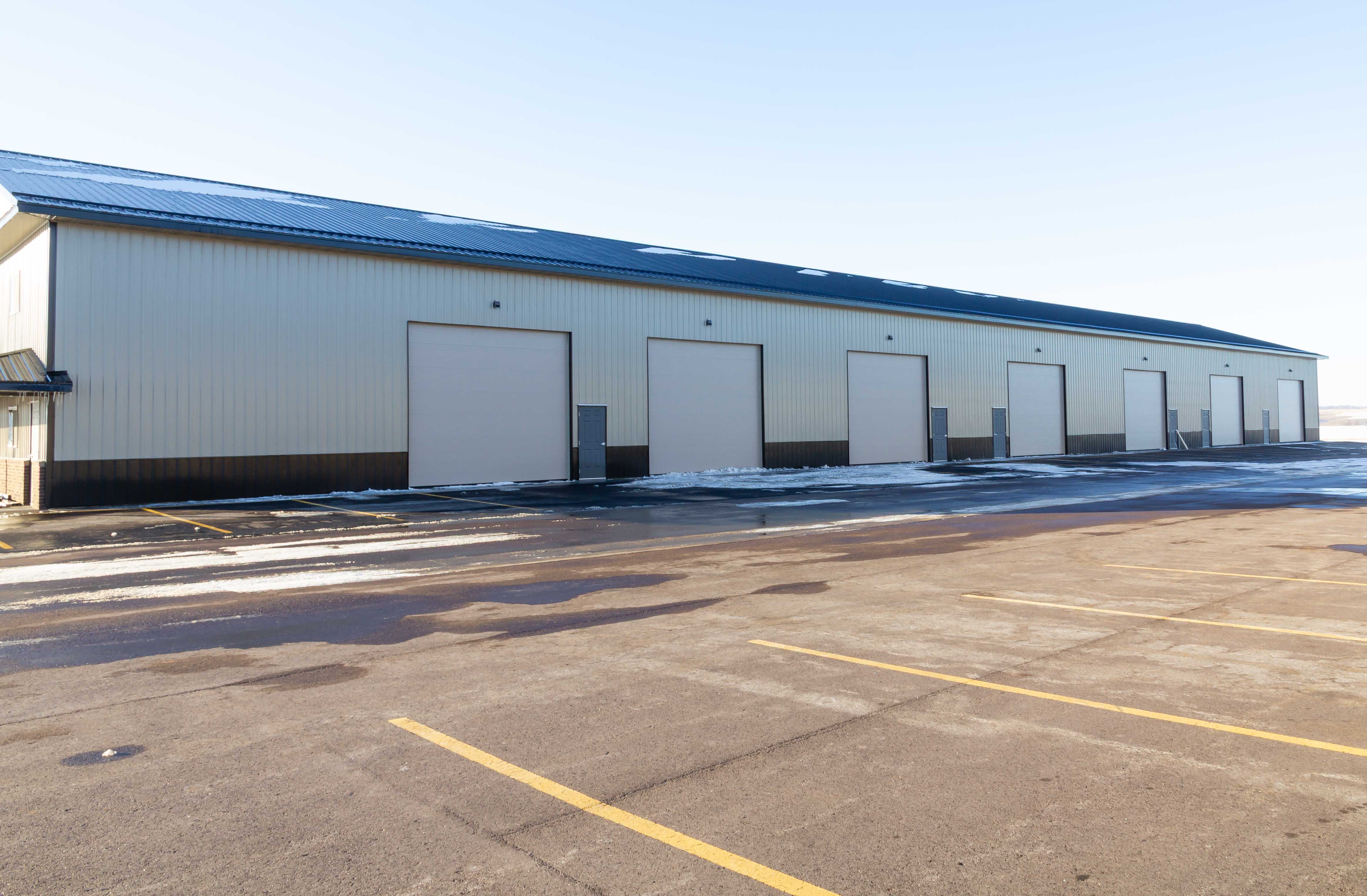 Side wall of a steel shop condo building lined with white overhead garage doors facing a striped parking lot