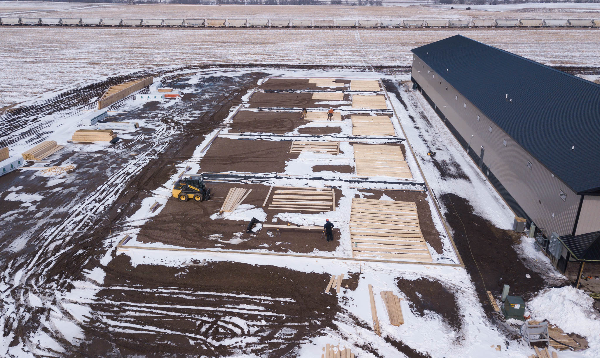 Aerial view of winter foundation work for new shop condos, with lumber stacks and a skid steer beside a finished building
