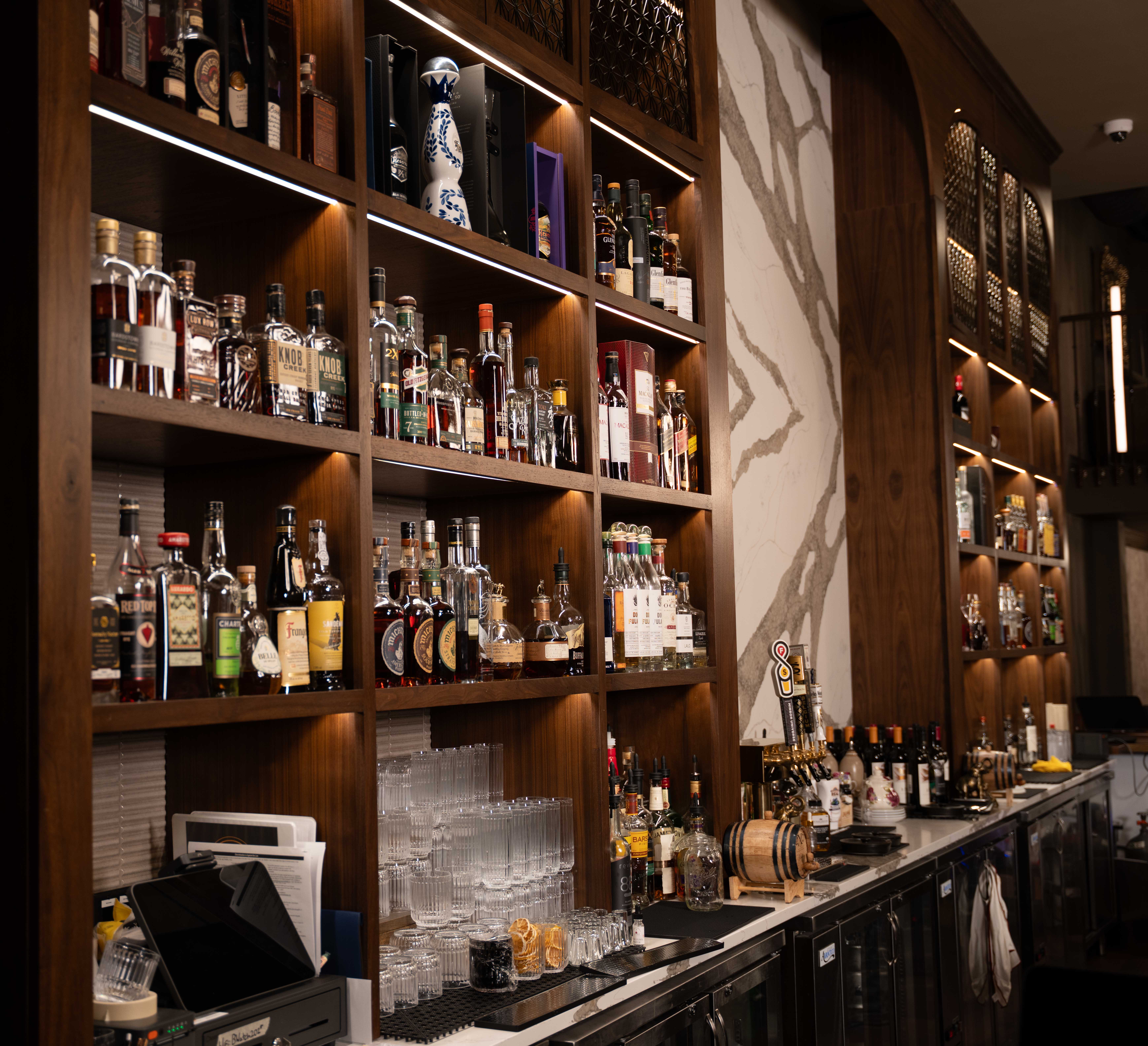 Backlit walnut shelving stocked with whiskey and spirits bottles beside a white marble panel behind the bar