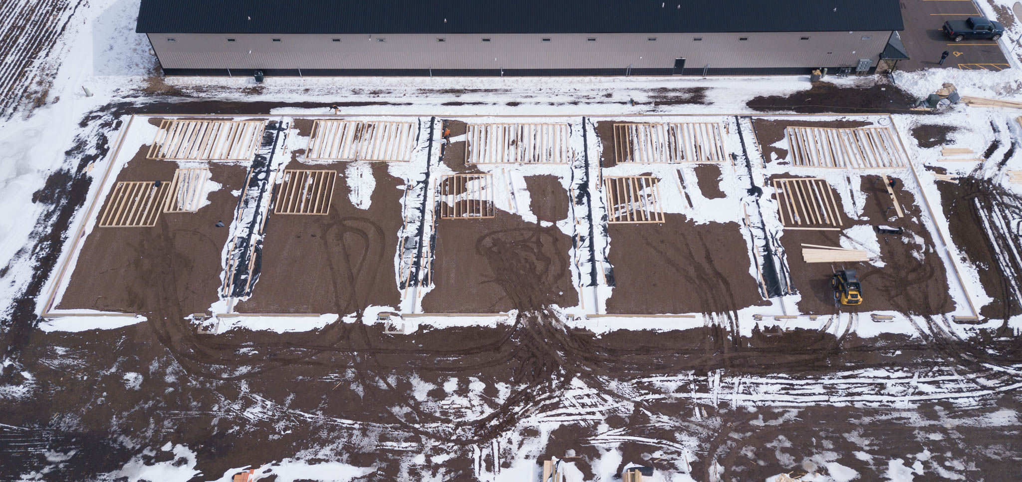 Overhead view of five foundation pads framed with lumber in snowy ground behind a finished shop condo building