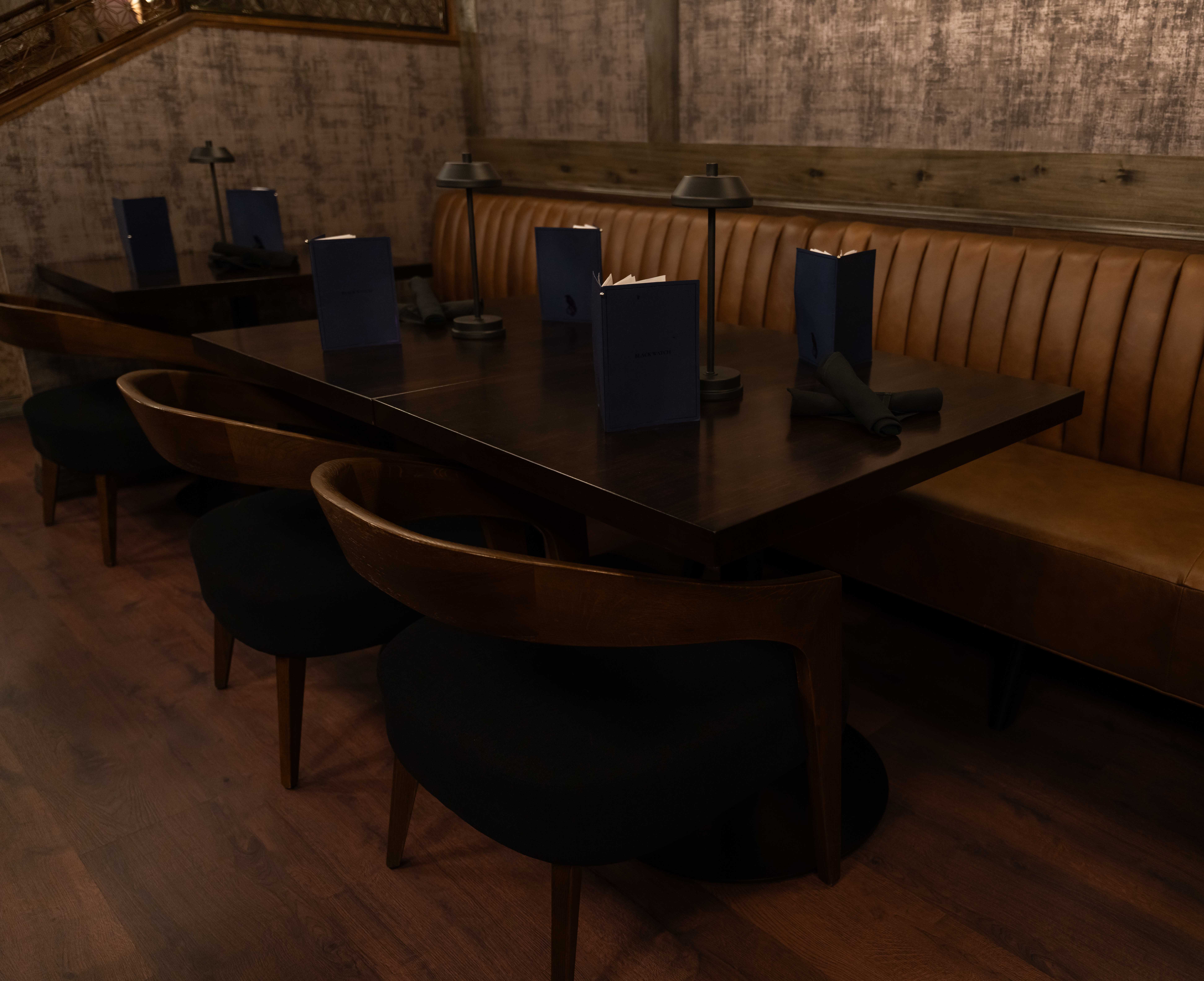 Dark wood dining tables with curved wood chairs and a tufted cognac leather banquette under brass table lamps