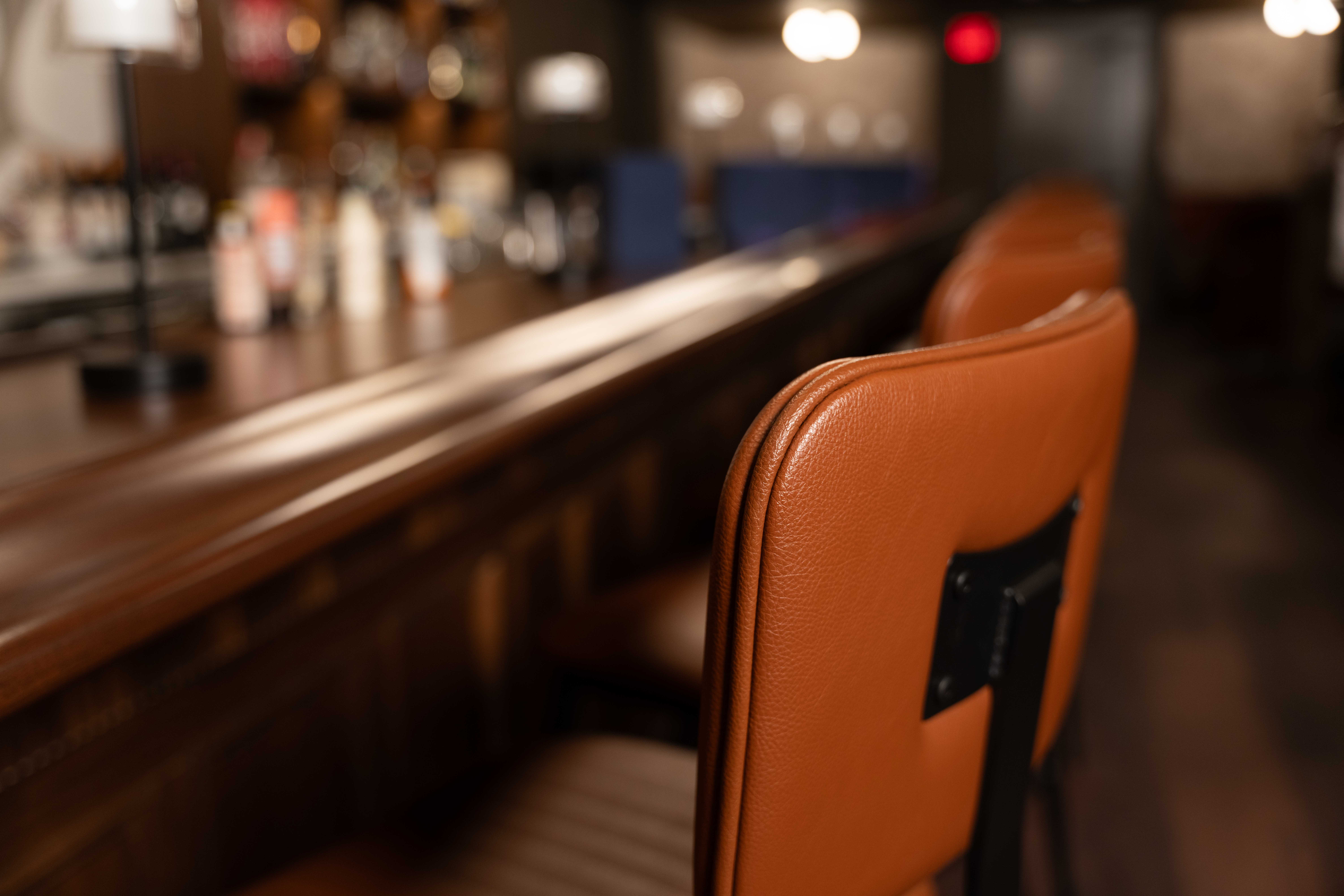 Close view of a cognac leather bar stool beside a polished dark wood bar rail with warm lighting beyond
