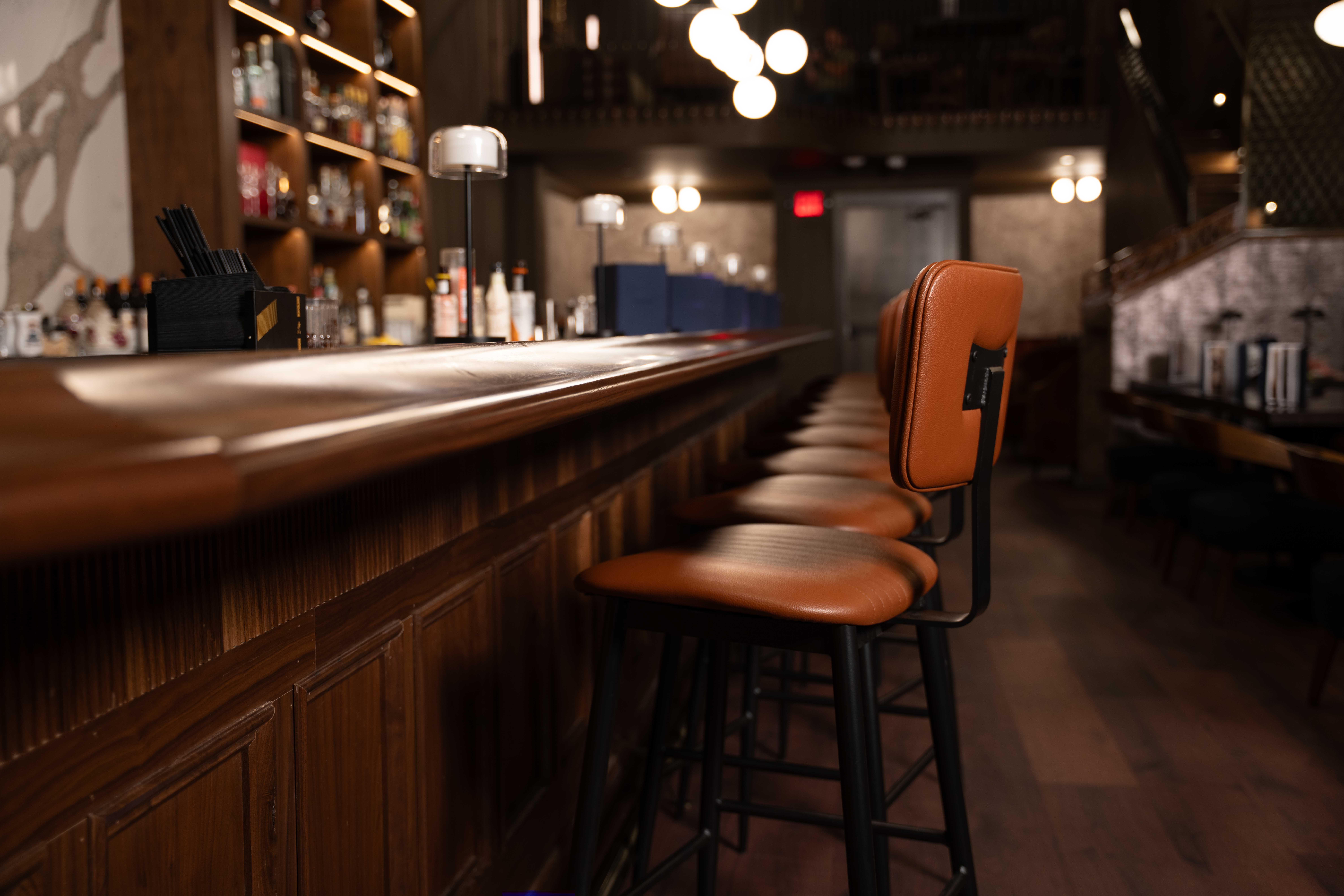 Leather-backed bar stools lined along a dark wood bar with liquor shelves and warm lighting in the background