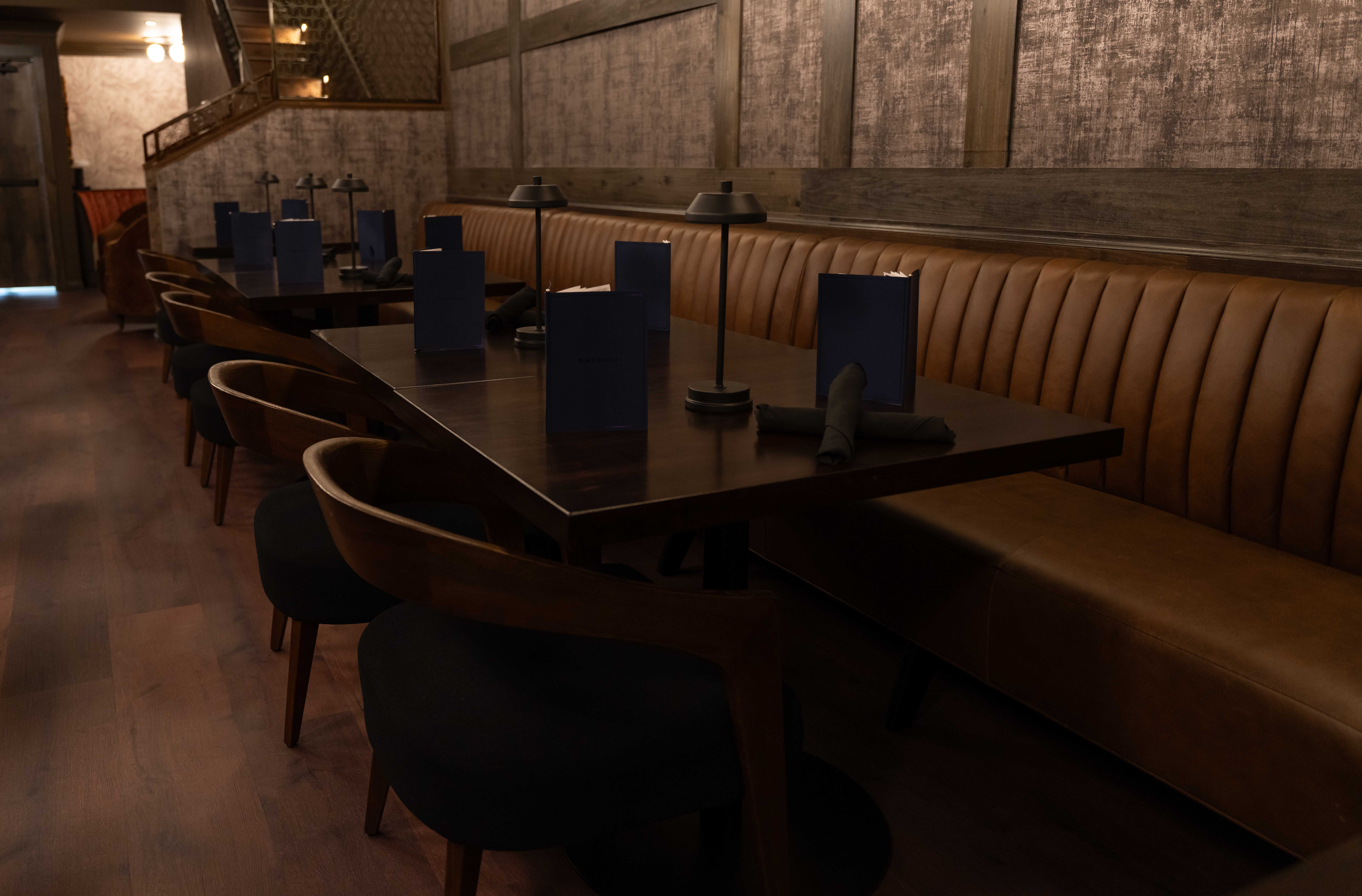 Row of dark wood tables with navy menus along a long channel-tufted leather banquette in a dim speakeasy