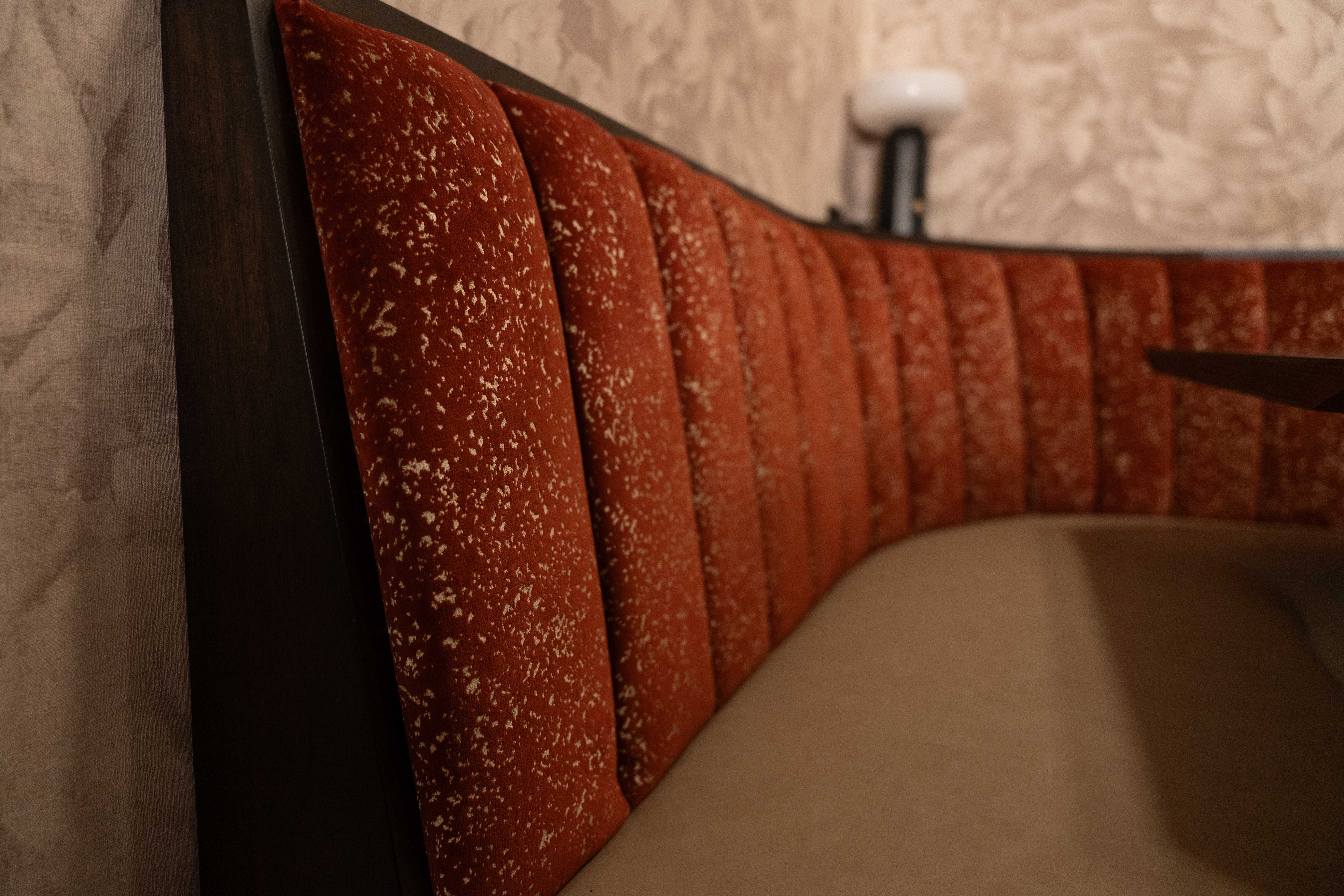 Close detail of a curved booth with rust-red channel-tufted velvet back and tan cushion against floral wallpaper
