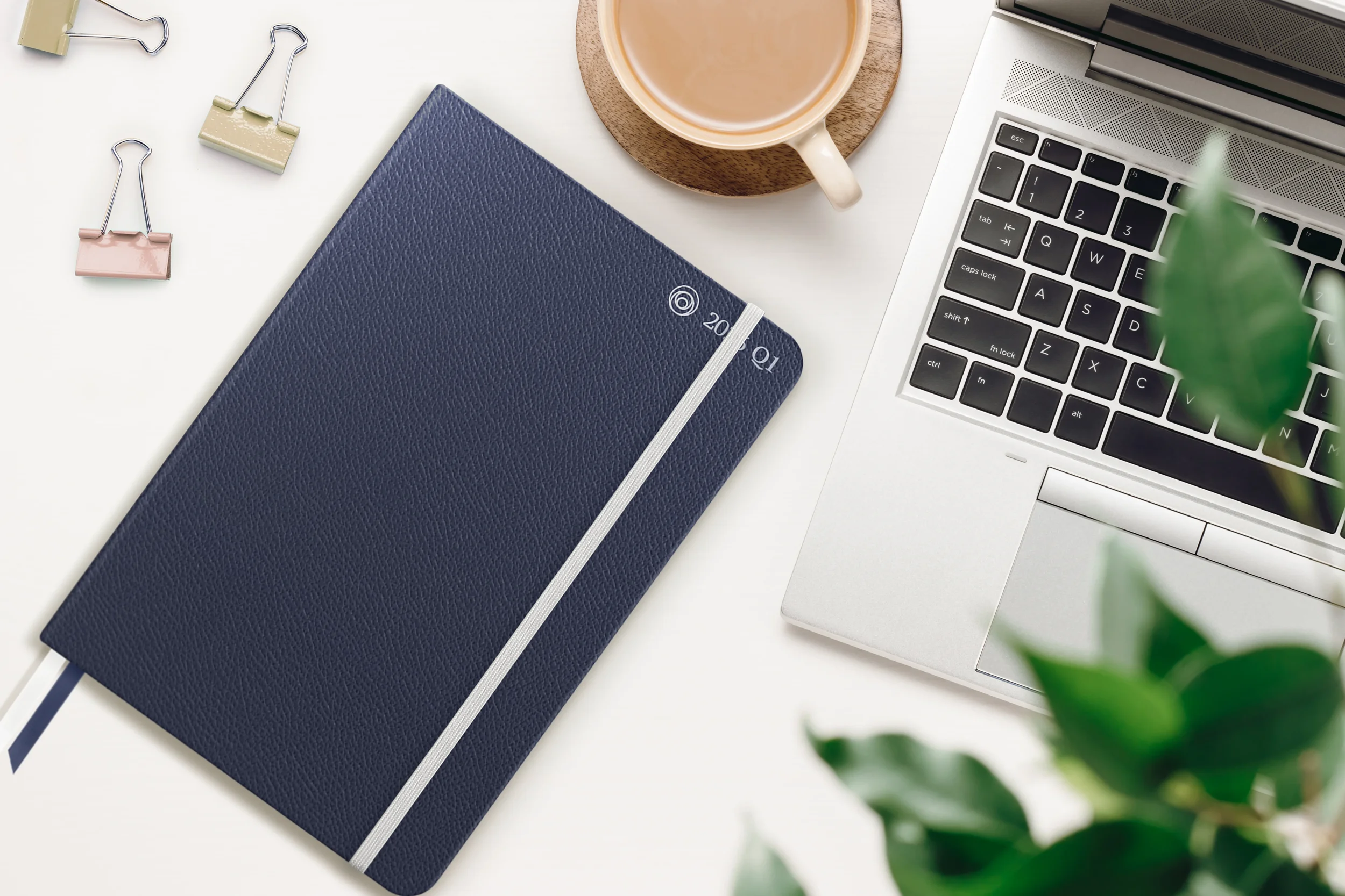 Workspace with a silver laptop keyboard, a navy blue 2021 Q1 planner with a white elastic band, a beige cup of coffee on a wooden coaster, three binder clips in pastel shades, and some green plant leaves partially visible.