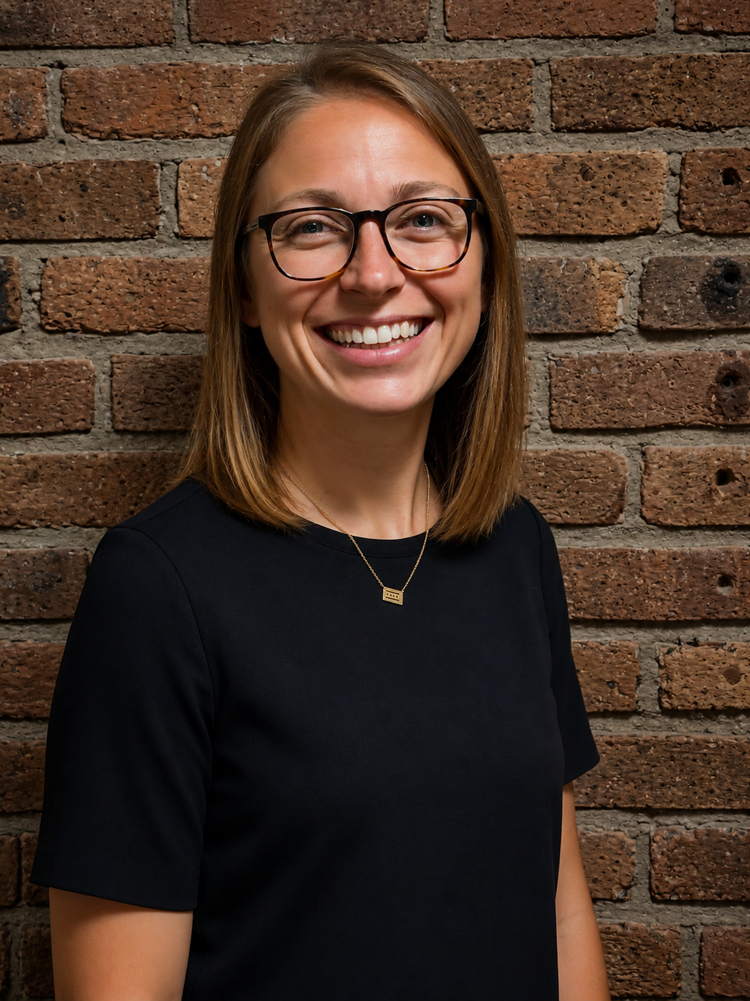 Allie Fish smiling in glasses and a black top against a brick wall, teen and family online therapist at Grow Wellness Group