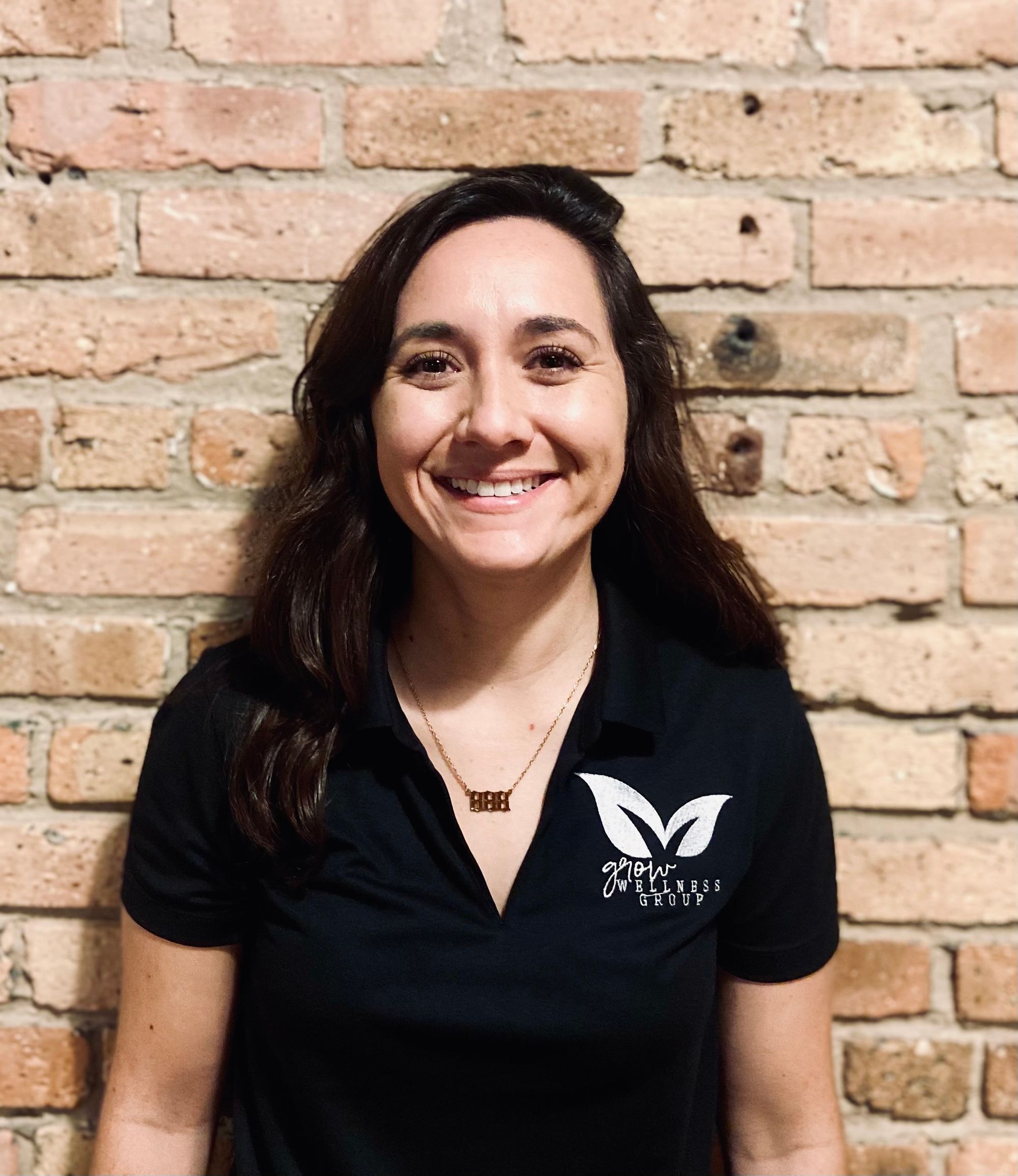 Christina Schrader smiling in a black Grow Wellness Group polo against a brick wall, sport psychology telehealth counselor