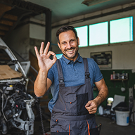 Smiling mechanic in overalls making an OK sign in a garage with car parts in the background.