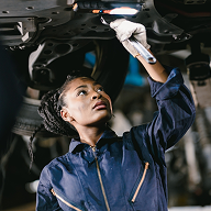 Female mechanic in blue coveralls working underneath a vehicle with a wrench.