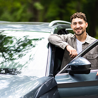 Smiling man leaning on the open driver's side window of a gray car with greenery in the background.