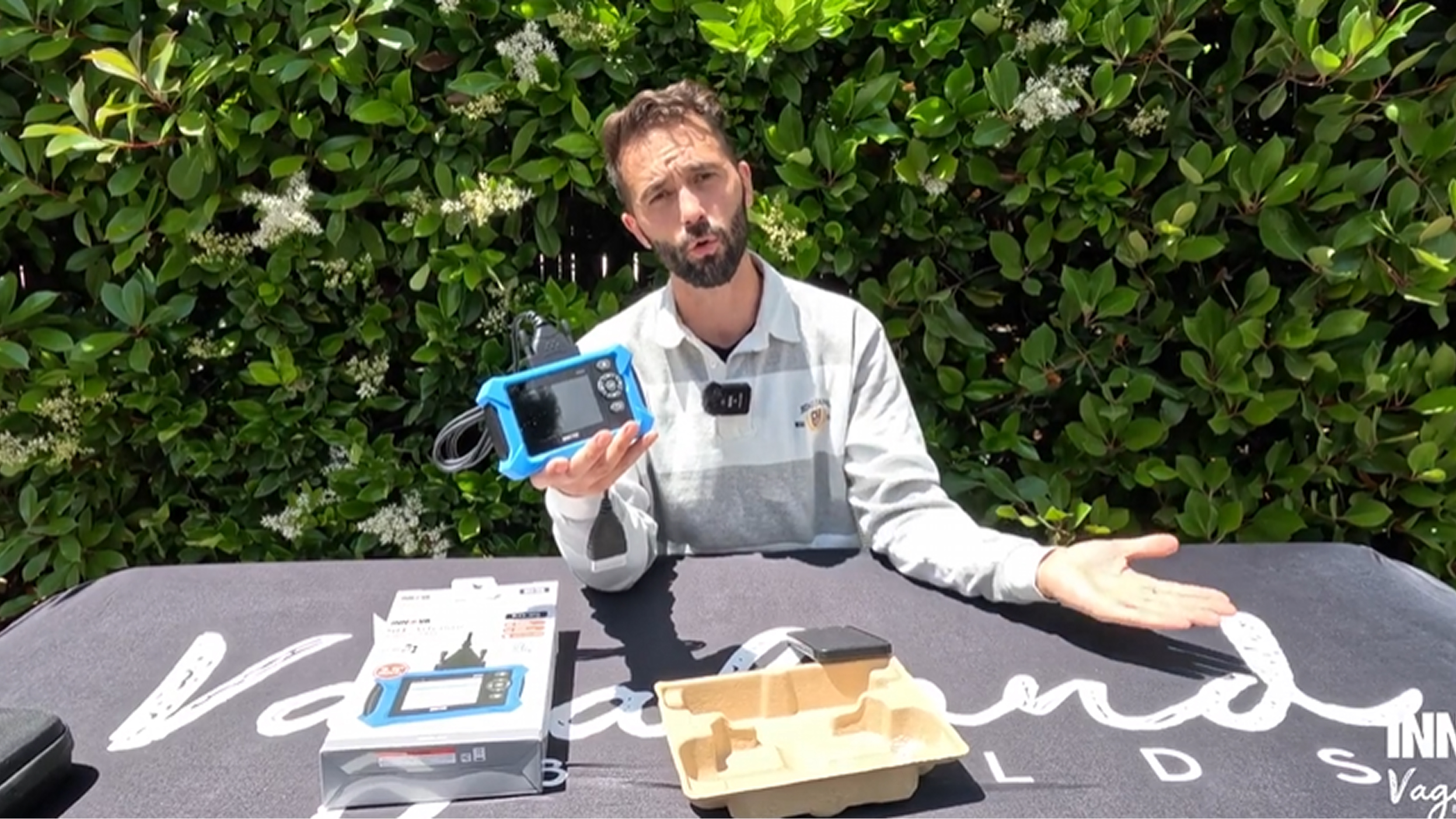 Man with a beard seated outdoors at a table, holding a blue electronic device with cables and gesturing with his other hand, with packaging and greenery in the background.
