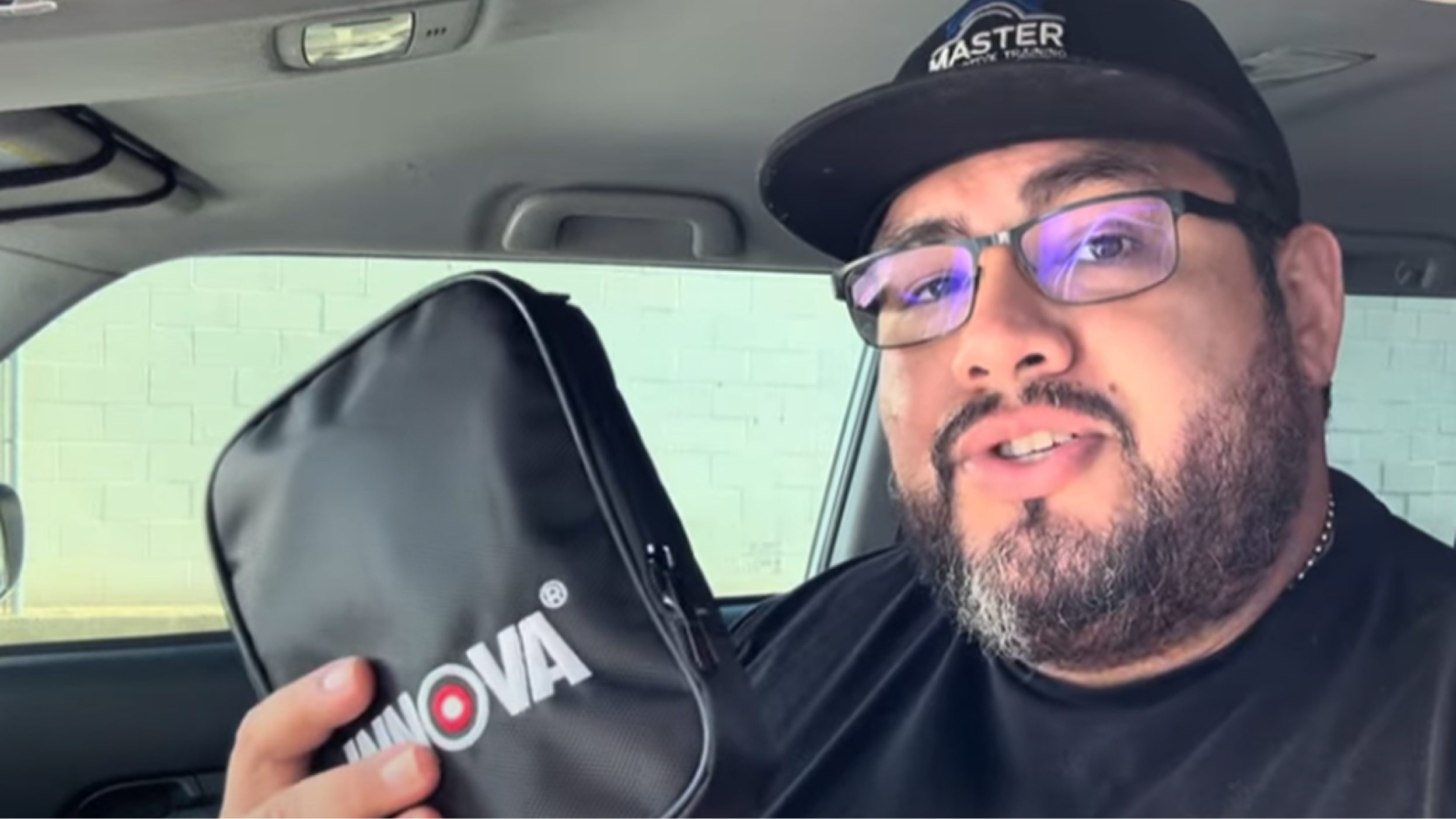 Man with glasses and a cap holding a black zippered pouch labeled 'INNOVA' inside a car.