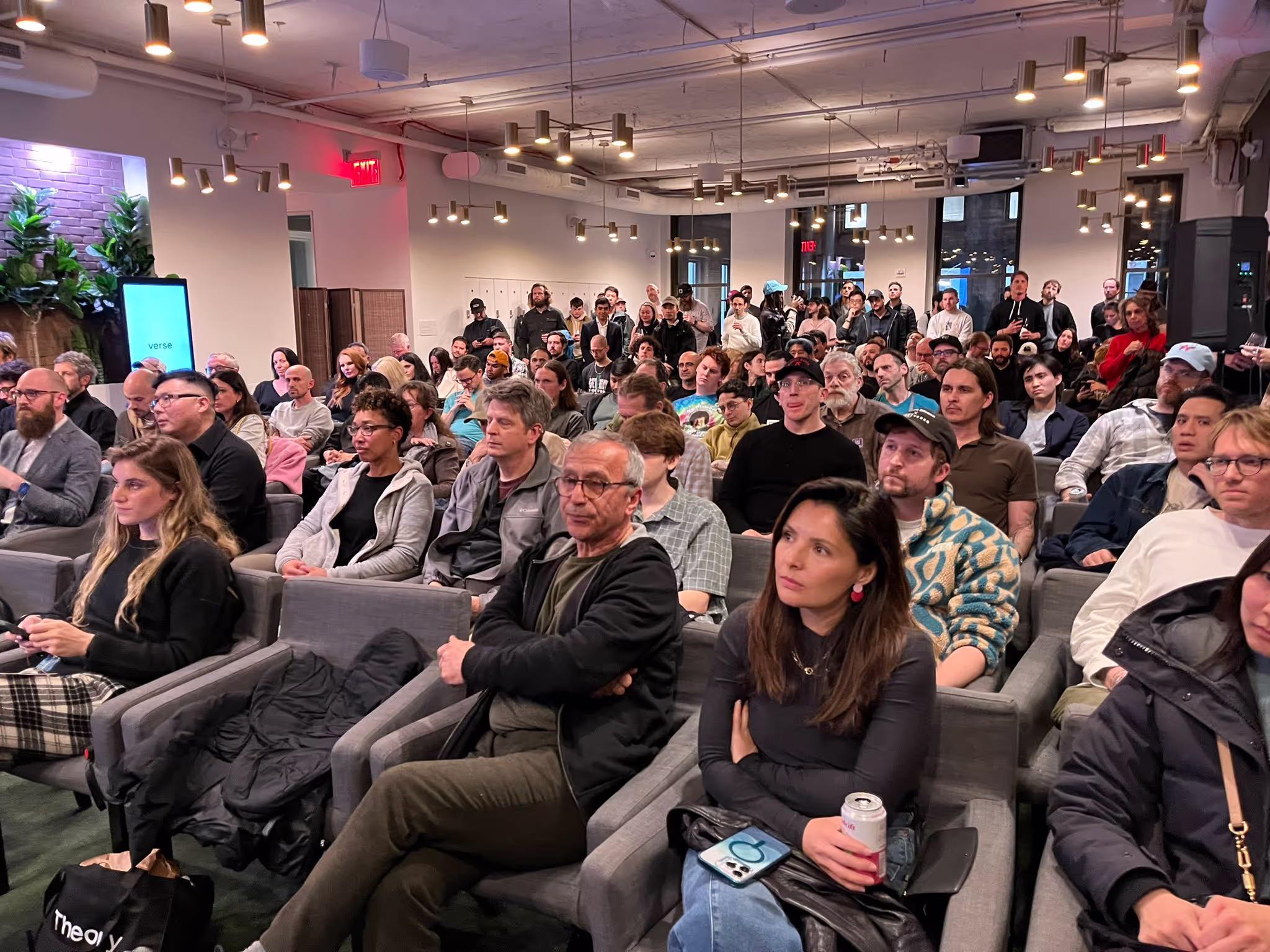 Crowd seated attentively in a modern room with soft lighting and plants on the left wall.