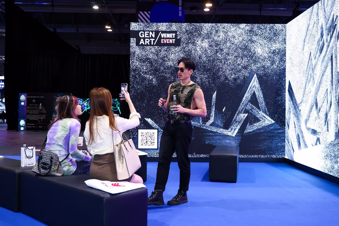 Two women sitting and taking a photo of a man posing with a bottle at a digital art event.