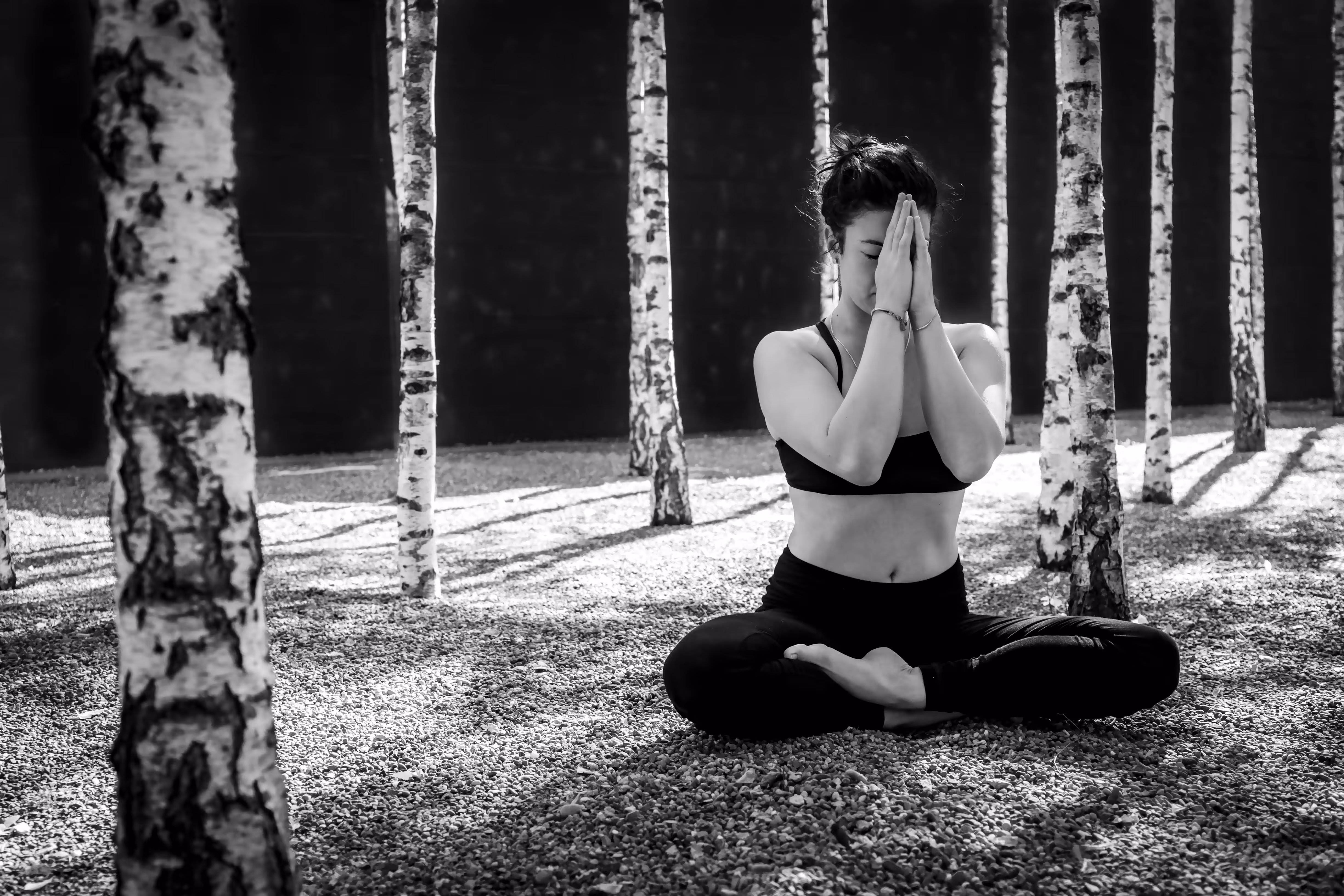 Woman in black sportswear sitting cross-legged meditating among birch trees on a gravel ground in black and white.