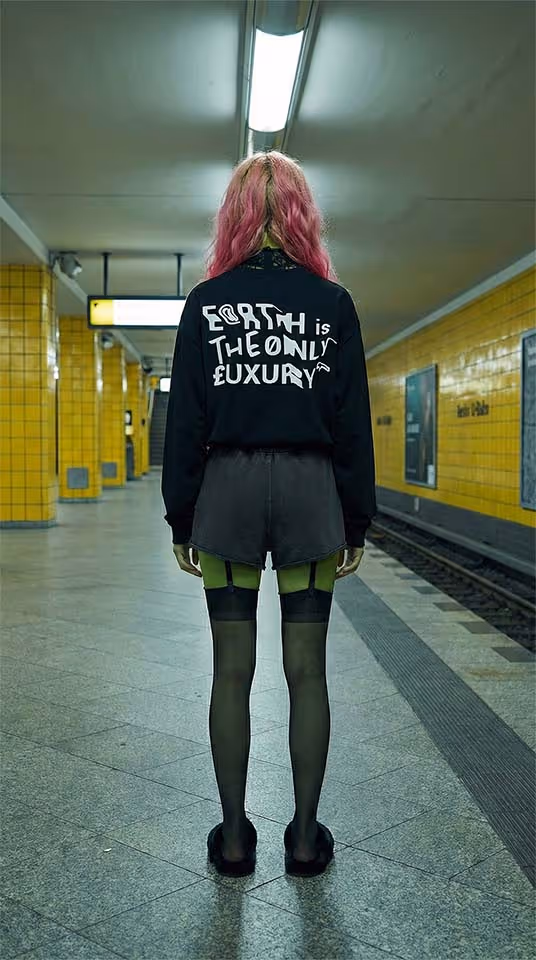 Person with pink hair standing in a subway station wearing a black sweatshirt with 'Earth is the only luxury' written on the back, black shorts, and green and black tights.