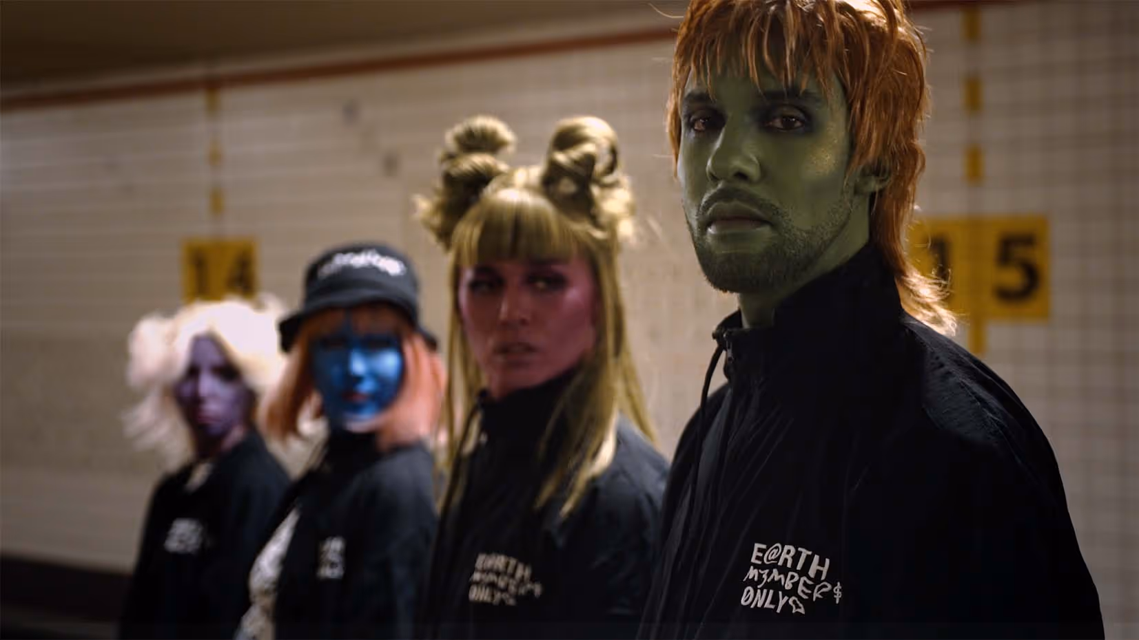 Four people standing in a line in a subway station, each with different colorful face paint and wearing black jackets with text.