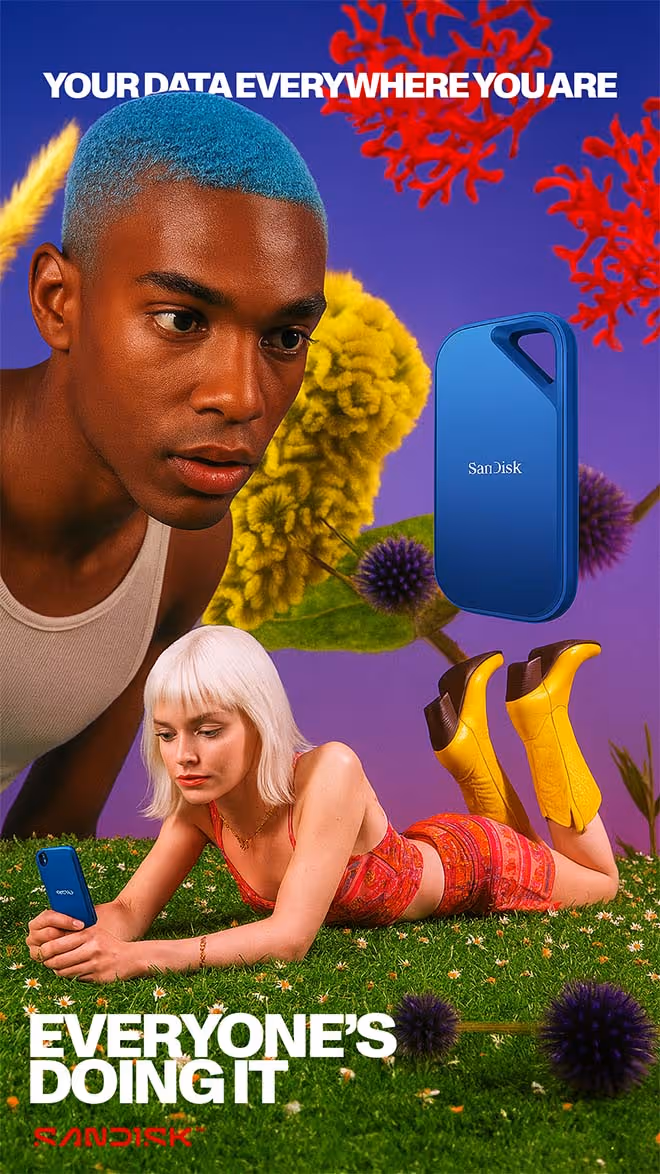 Advertisement showing a man with bright blue hair and a woman in a red outfit lying on grass using a smartphone, with a blue SanDisk device floating and text reading 'Your data everywhere you are' and 'Everyone's doing it'.