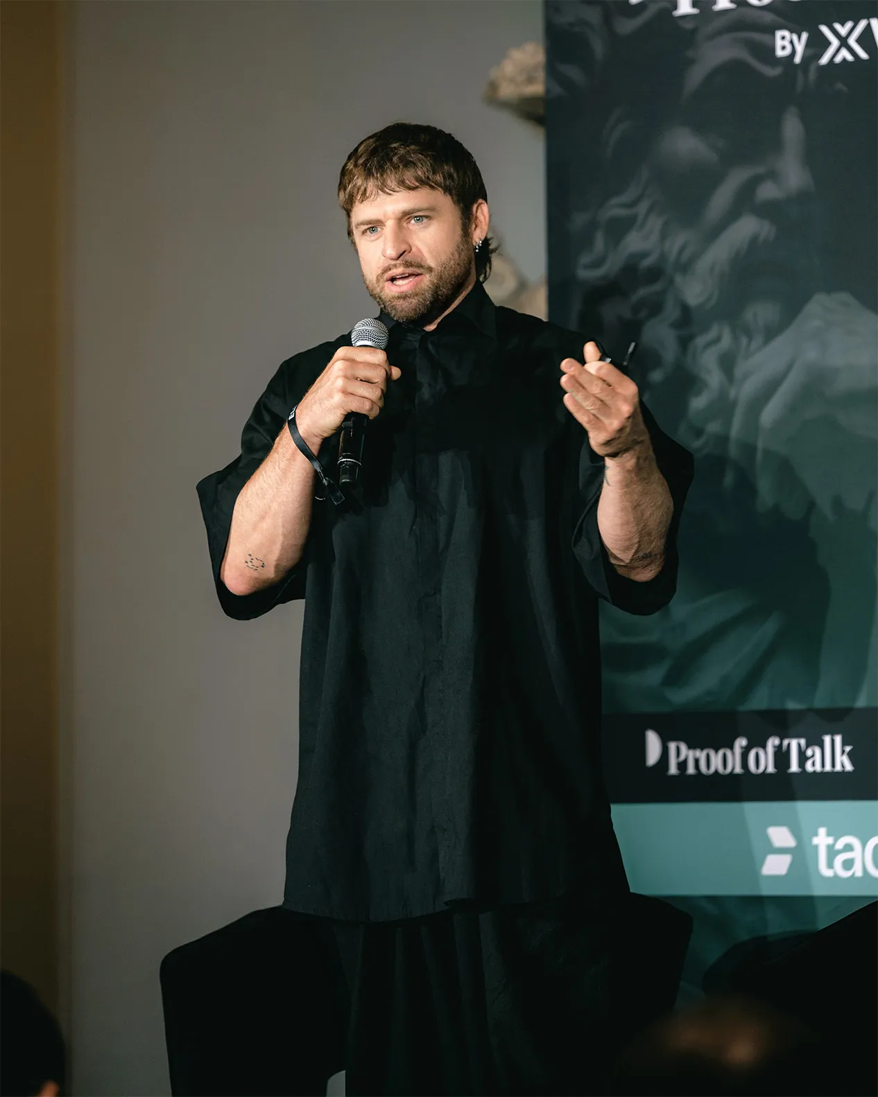 Man with beard speaking into microphone wearing loose black shirt, with a banner in the background.
