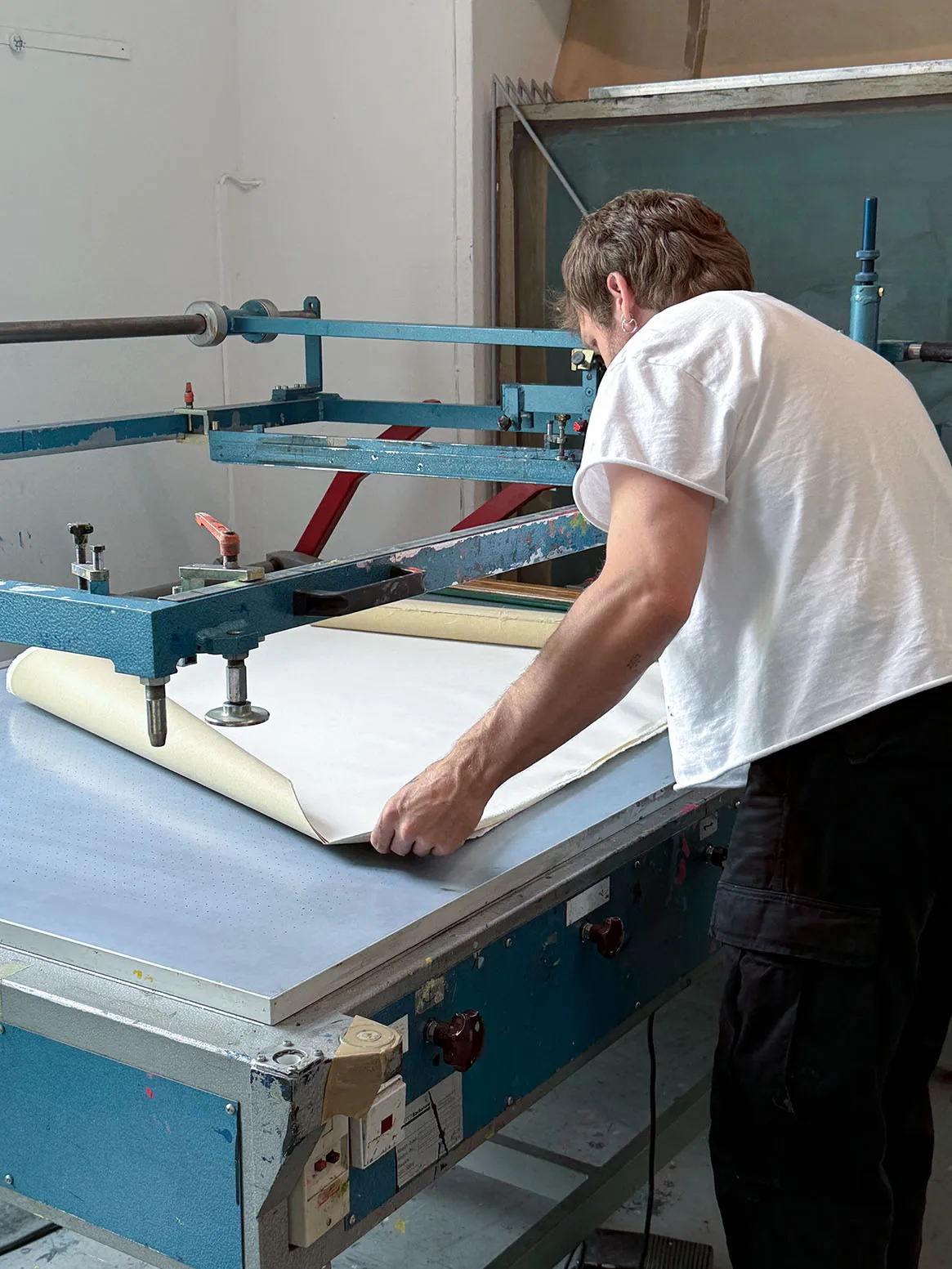 Person in white shirt handling large sheet on blue industrial screen printing machine.