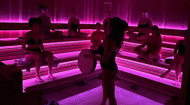 Small group of women sit in a sauna, dimly lit by pink light 