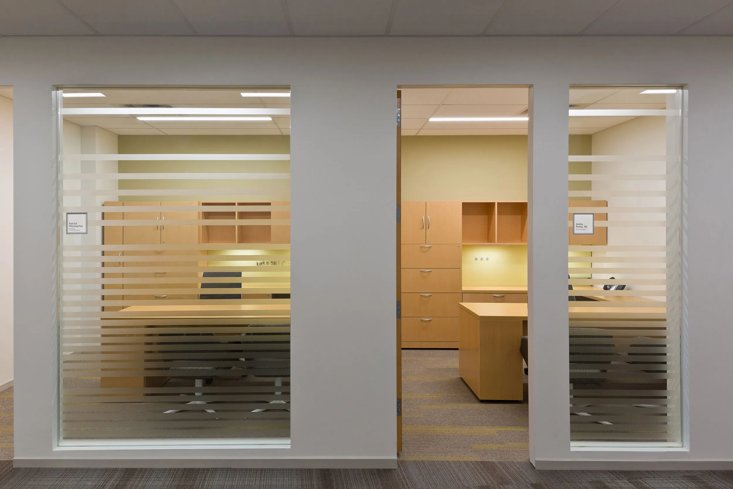 Modern office space with two workstations, wooden desks, storage cabinets, and frosted glass walls with horizontal stripes.