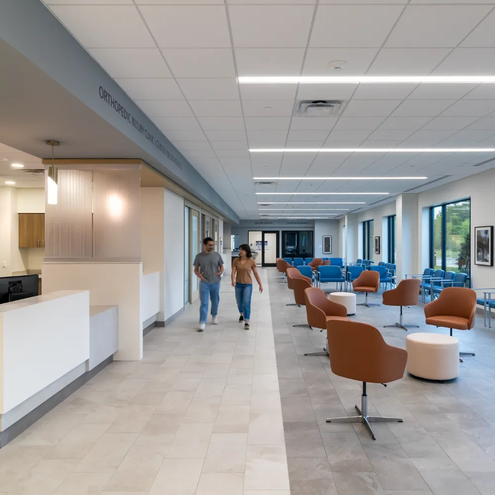 Modern orthopedic clinic waiting area with brown and blue chairs, large windows, and two people walking down the corridor.