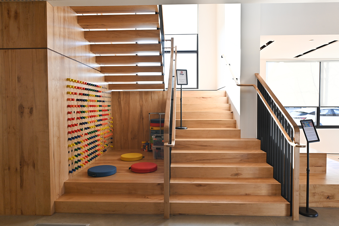 Modern wooden staircase with colorful pegboard game and padded floor cushions in a bright, open space.