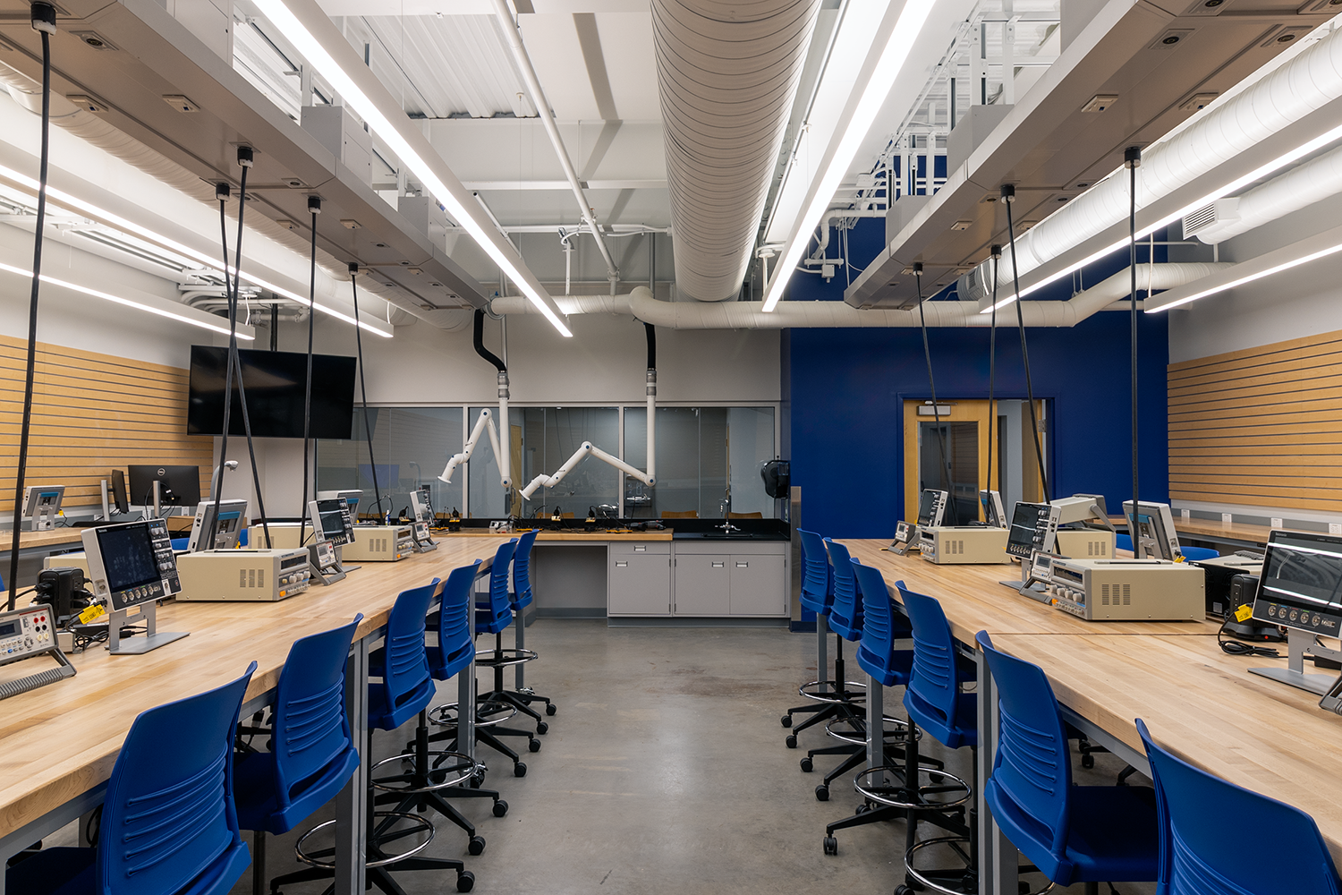 Modern laboratory with rows of blue chairs along wooden workbenches equipped with electronic testing equipment and monitors.