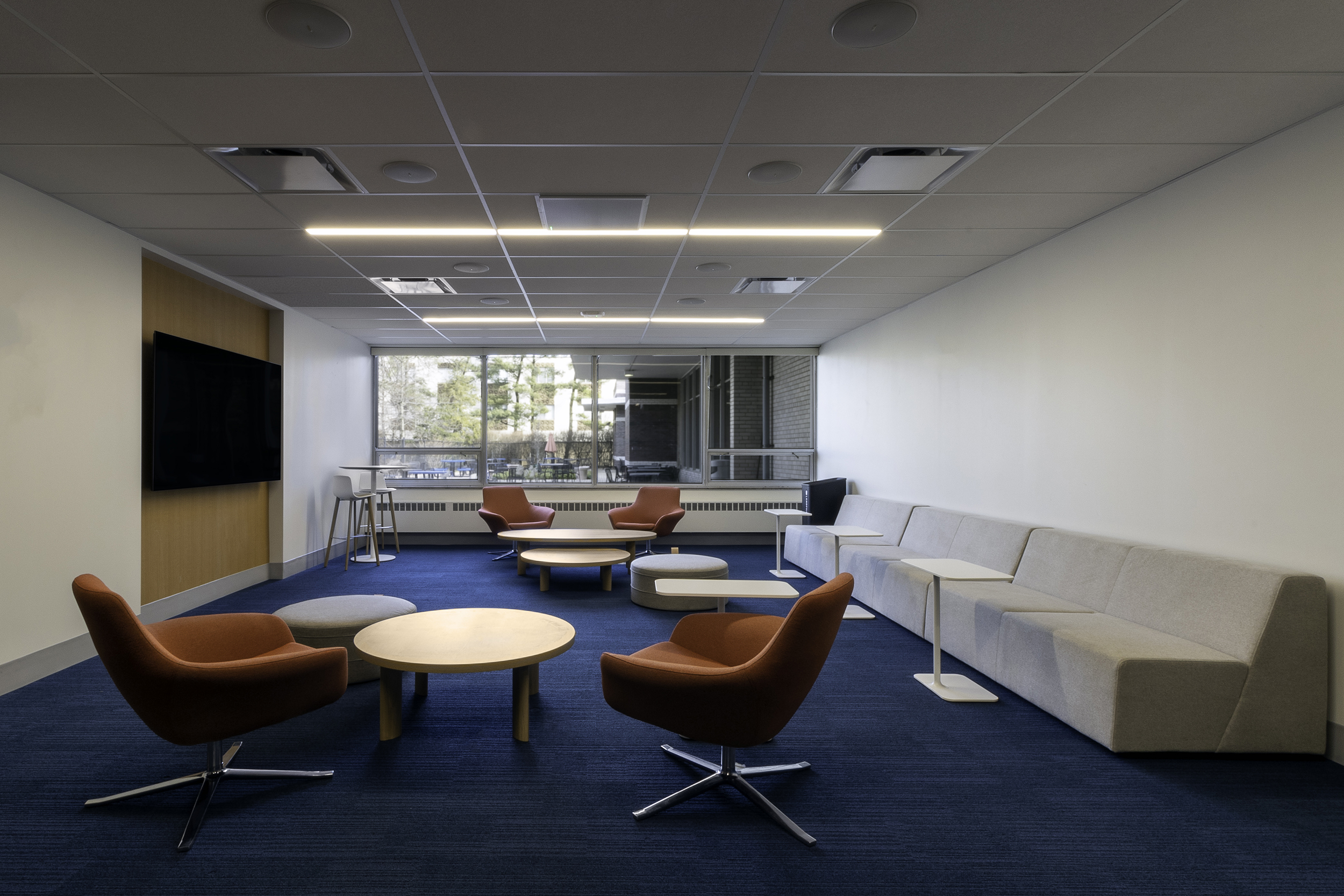 Modern office lounge with orange swivel chairs, grey sofa, round wooden tables, and blue carpet under ceiling lights.