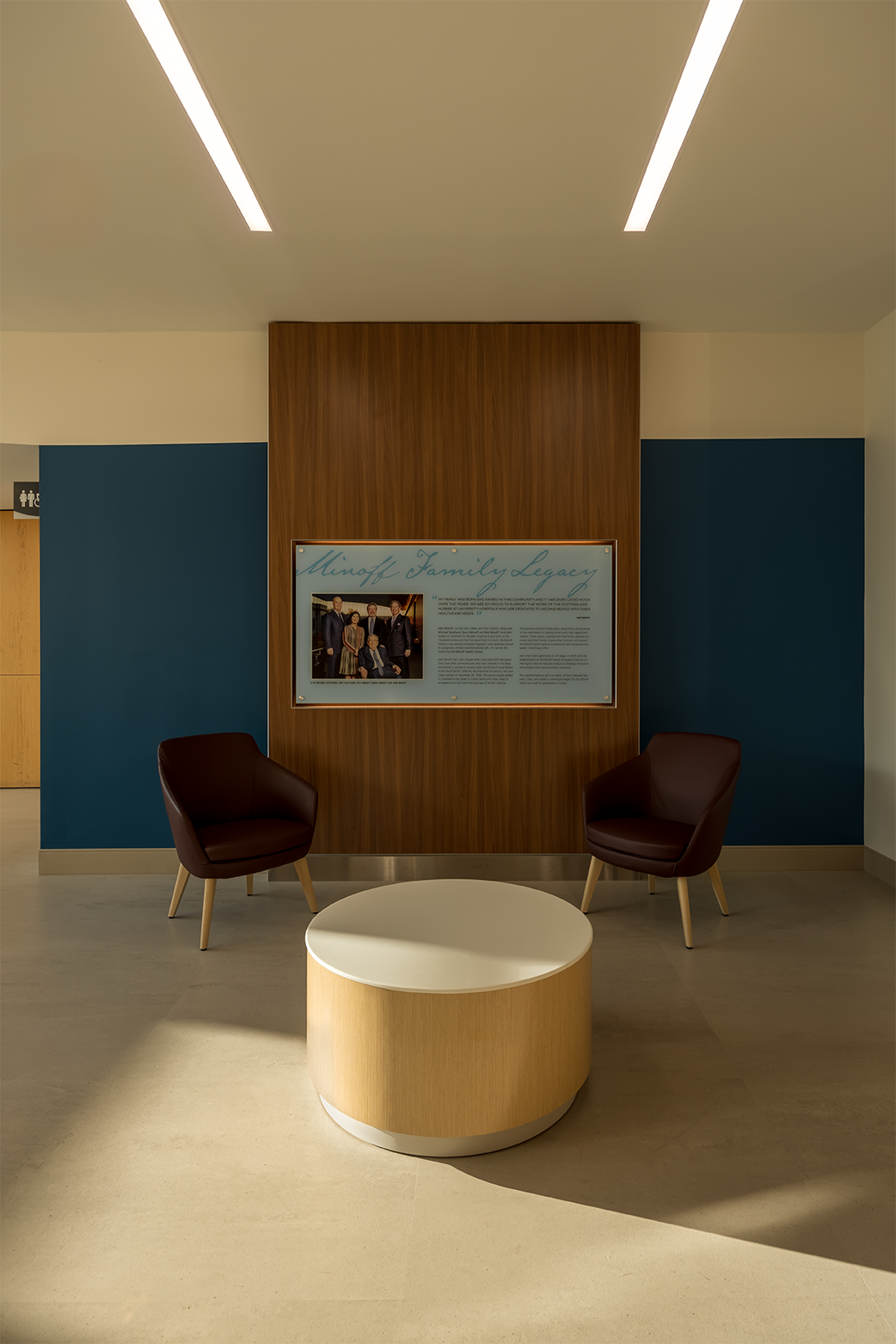 Modern office lounge with two women sitting on a brown sectional sofa working on laptops, two orange chairs, a round wooden table, whiteboards, and wall-mounted TVs.
