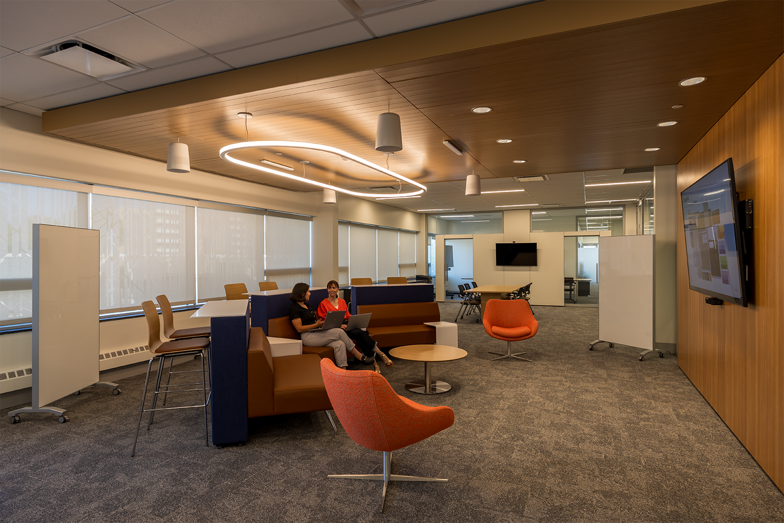 Modern office lounge area with two women sitting on a brown couch working on laptops, orange chairs, whiteboards, and large windows with blinds.