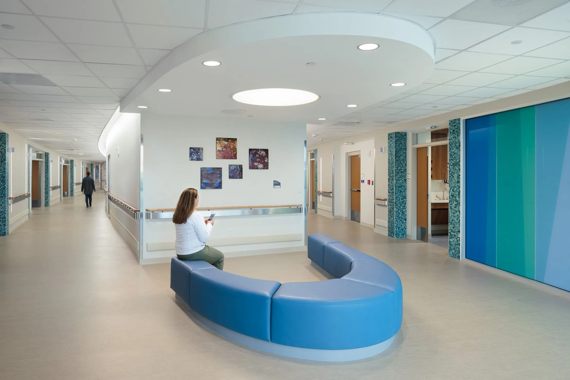 Modern hospital corridor with curved blue seating and a woman sitting on it looking at her phone.