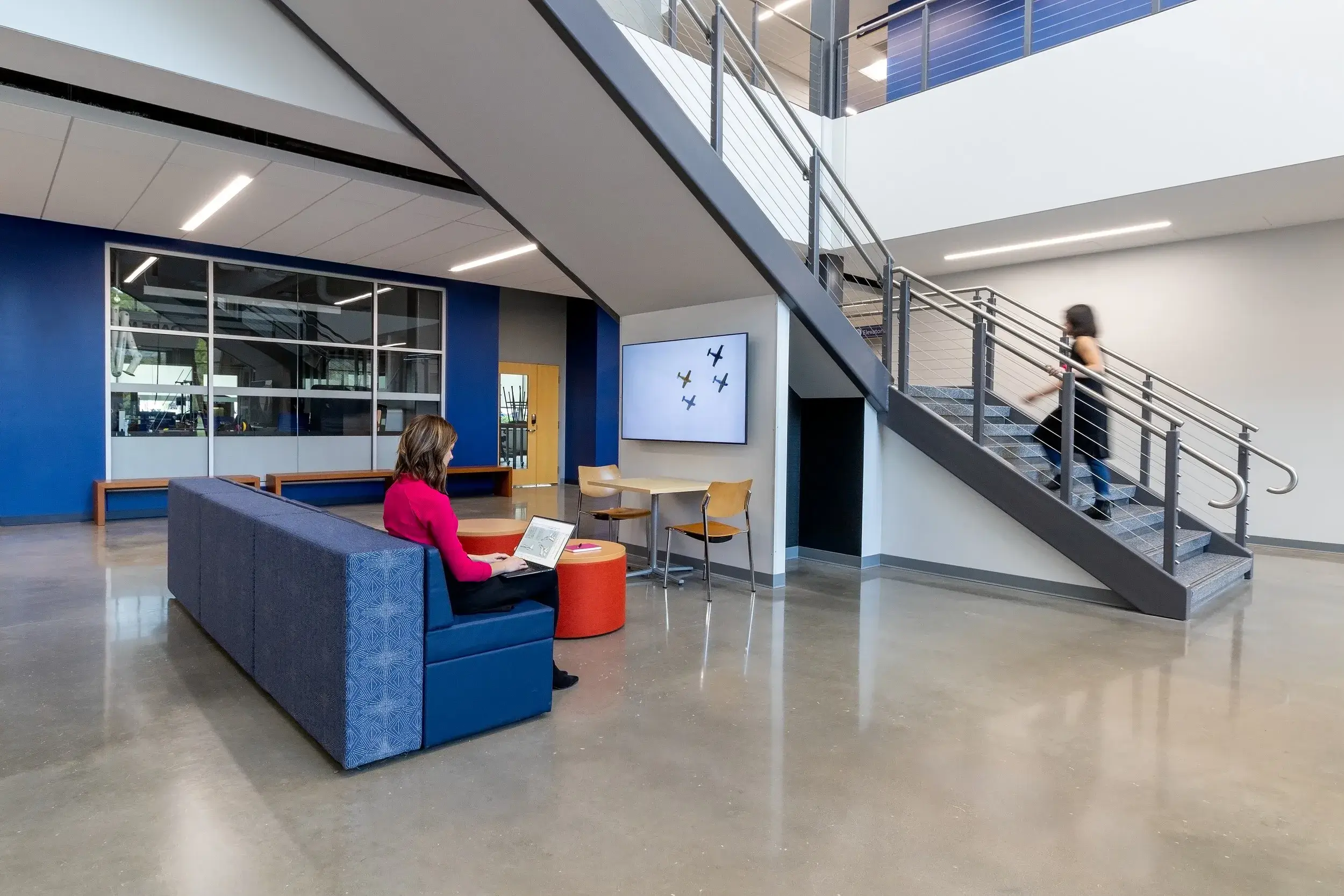 Modern indoor common area with a woman sitting on a blue couch using a laptop and another woman walking up a staircase.