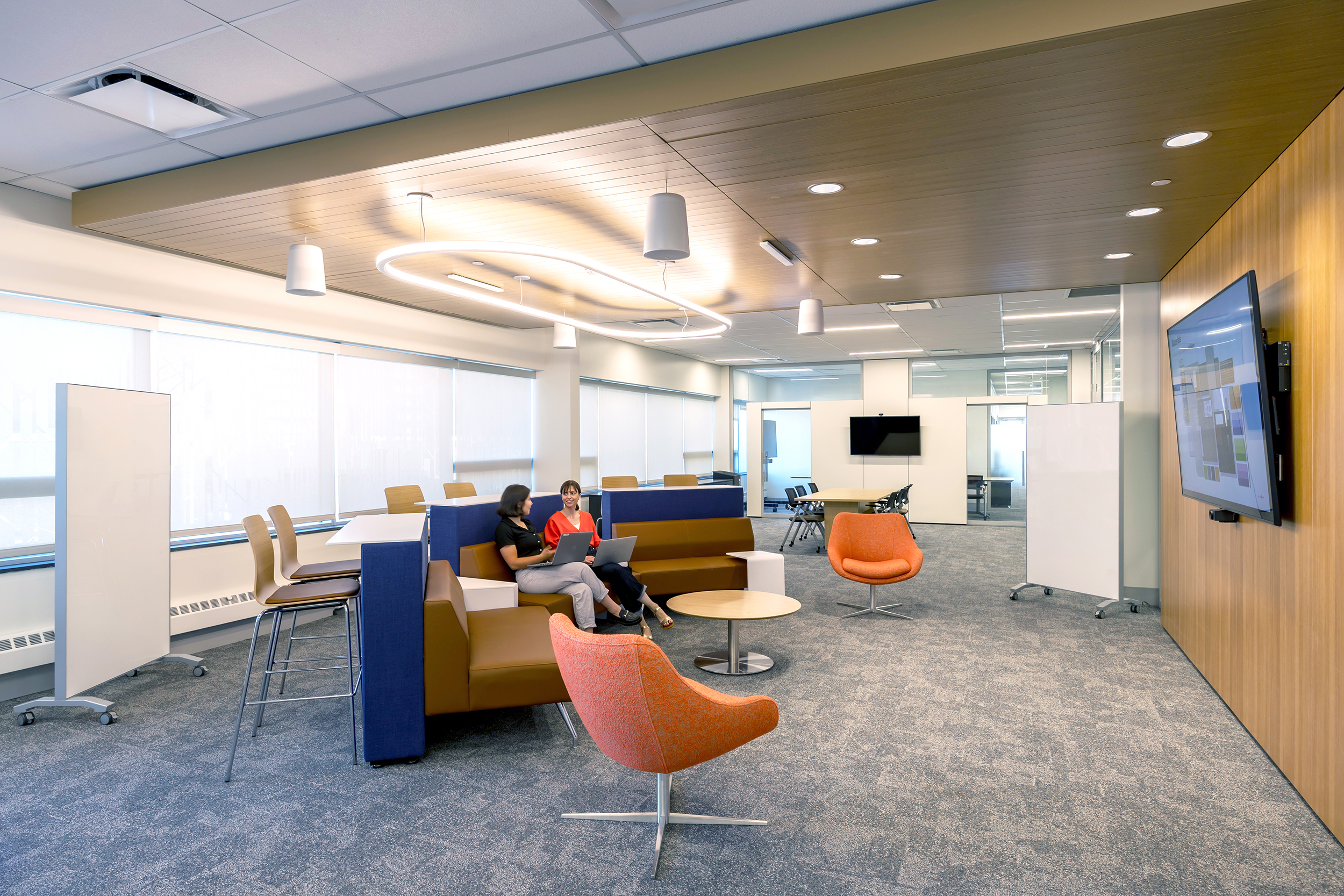 Modern office lounge area with two women sitting on a brown couch working on laptops, orange chairs, whiteboards, and large windows with blinds.