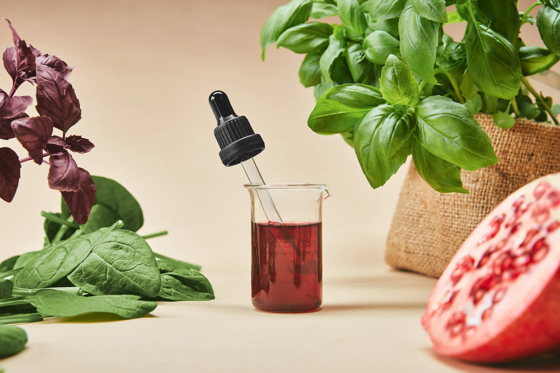 Dropper and beaker of red liquid surrounded by fresh basil, spinach, and pomegranate