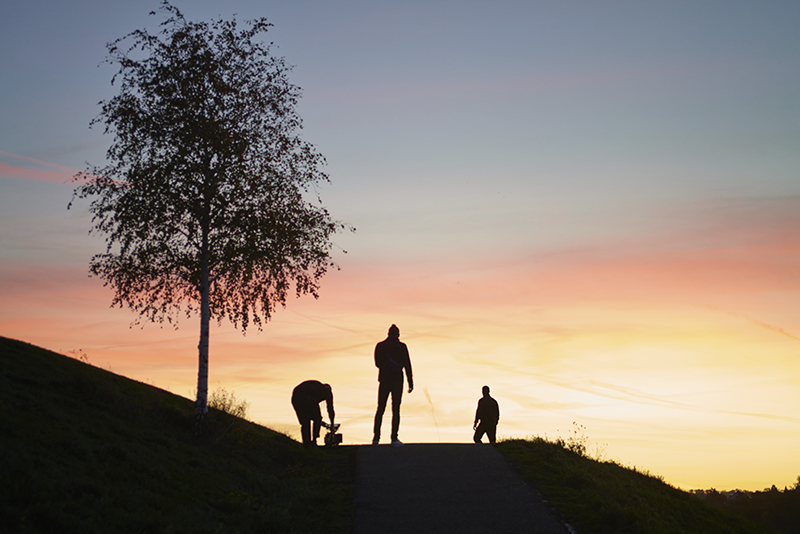 Ein Schwarz-Weiß-Design mit einer zentral platzierten Szene eines Filmsets, in der ein Kameramann auf einem Holzhocker sitzt und eine Kamera justiert, während das Team um ihn herum arbeitet. Darüber steht „WIR SIND“ mit funkelnden Symbolen, darunter die Begriffe „IDEEN“, „DETAILS“, „REALISATION“ und „KREATIV“.