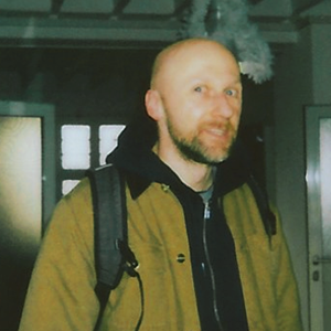 Ein Mann mit Glatze und Bart trägt einen braunen Mantel und einen Rucksack in einem Innenraum.