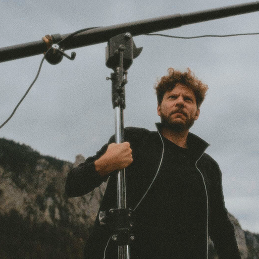 Ein Mann mit lockigem Haar und schwarzer Jacke hält eine große Stativstange, im Hintergrund Berge und bewölkter Himmel.