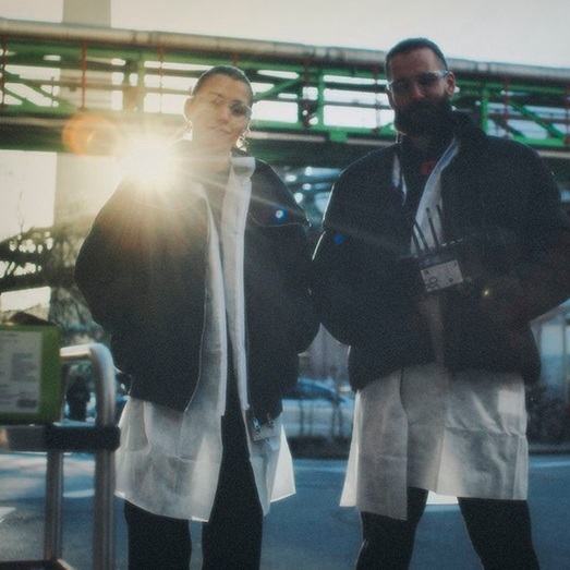 Zwei Personen mit medizinischer Schutzkleidung und Jacken, stehend im Freien bei hellem Sonnenlicht.