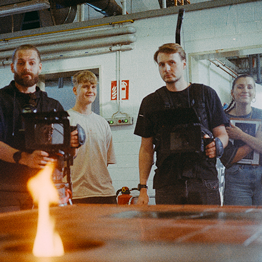 Vier junge Menschen beobachten eine Flamme in einer Werkstatt mit technischer Ausrüstung im Hintergrund.