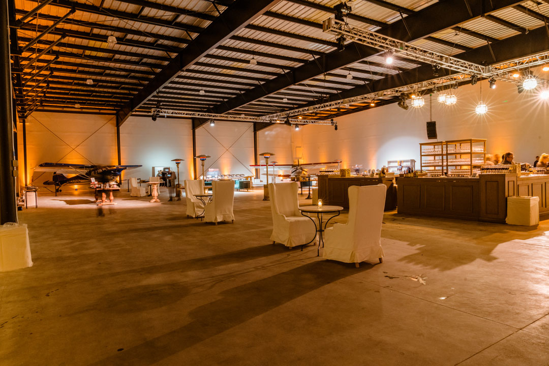 Empty hangar interior at The Executive Hangar with white slipcovered chairs, cocktail tables, long bar and a small plane