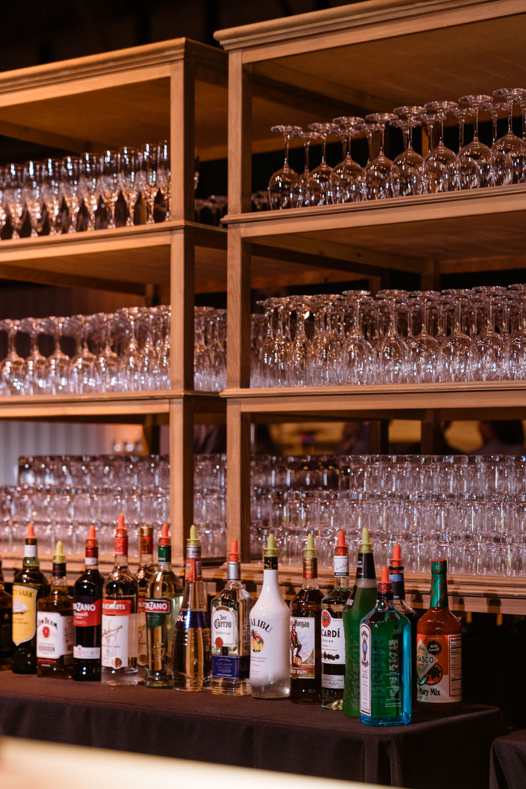 Back bar with wood shelves of stacked glassware and a row of liquor bottles on a black-draped table