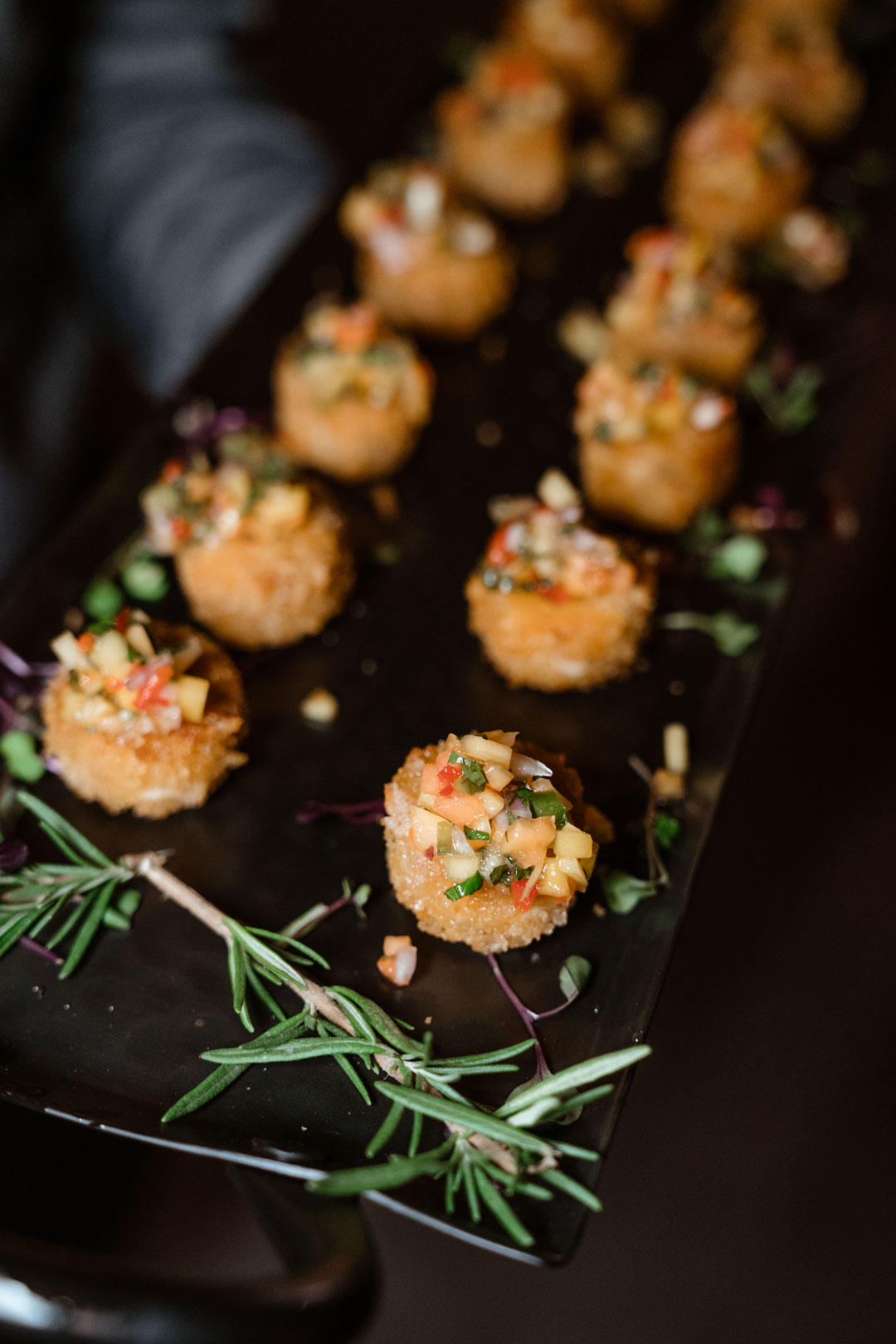 Crab cakes topped with diced fruit salsa on a black serving tray with rosemary