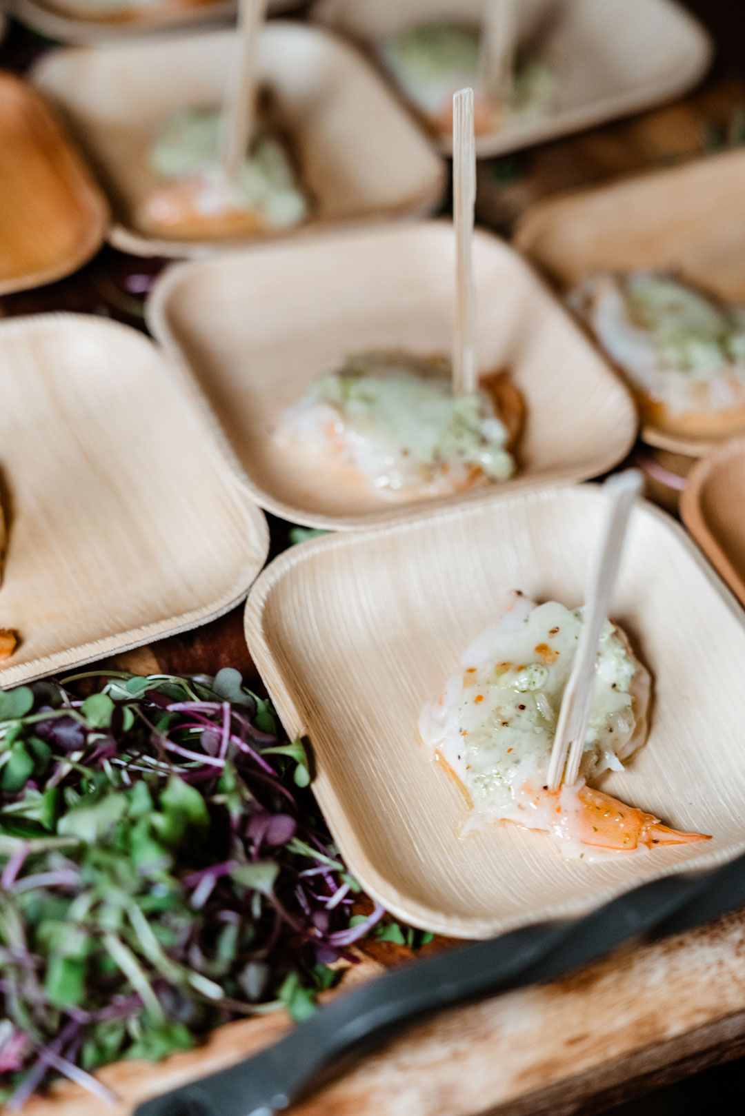 Shrimp mascarpone bites on skewers in square palm leaf plates beside purple microgreens